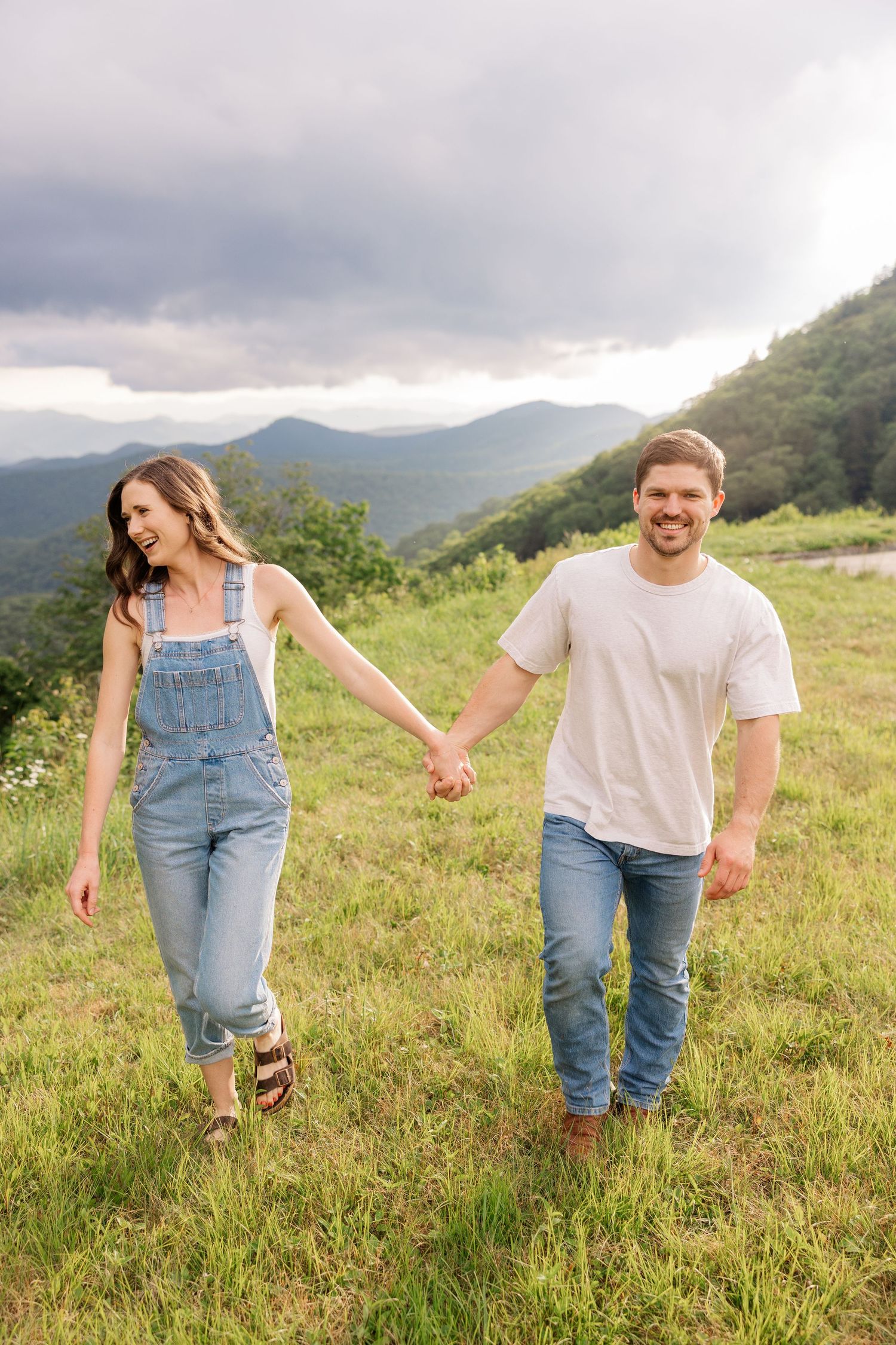 A couple walks hand in hand through a scenic mountain meadow with dramatic cloudy skies overhead.