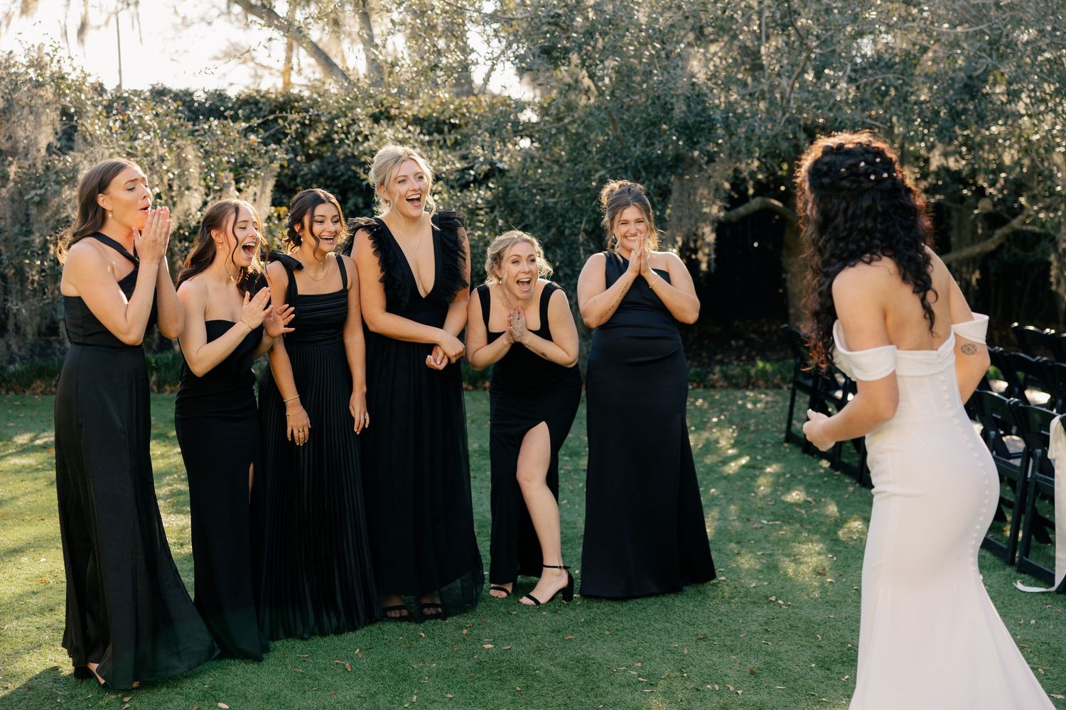Bridesmaids in black dresses react with joy to seeing bride in white wedding gown outdoors.