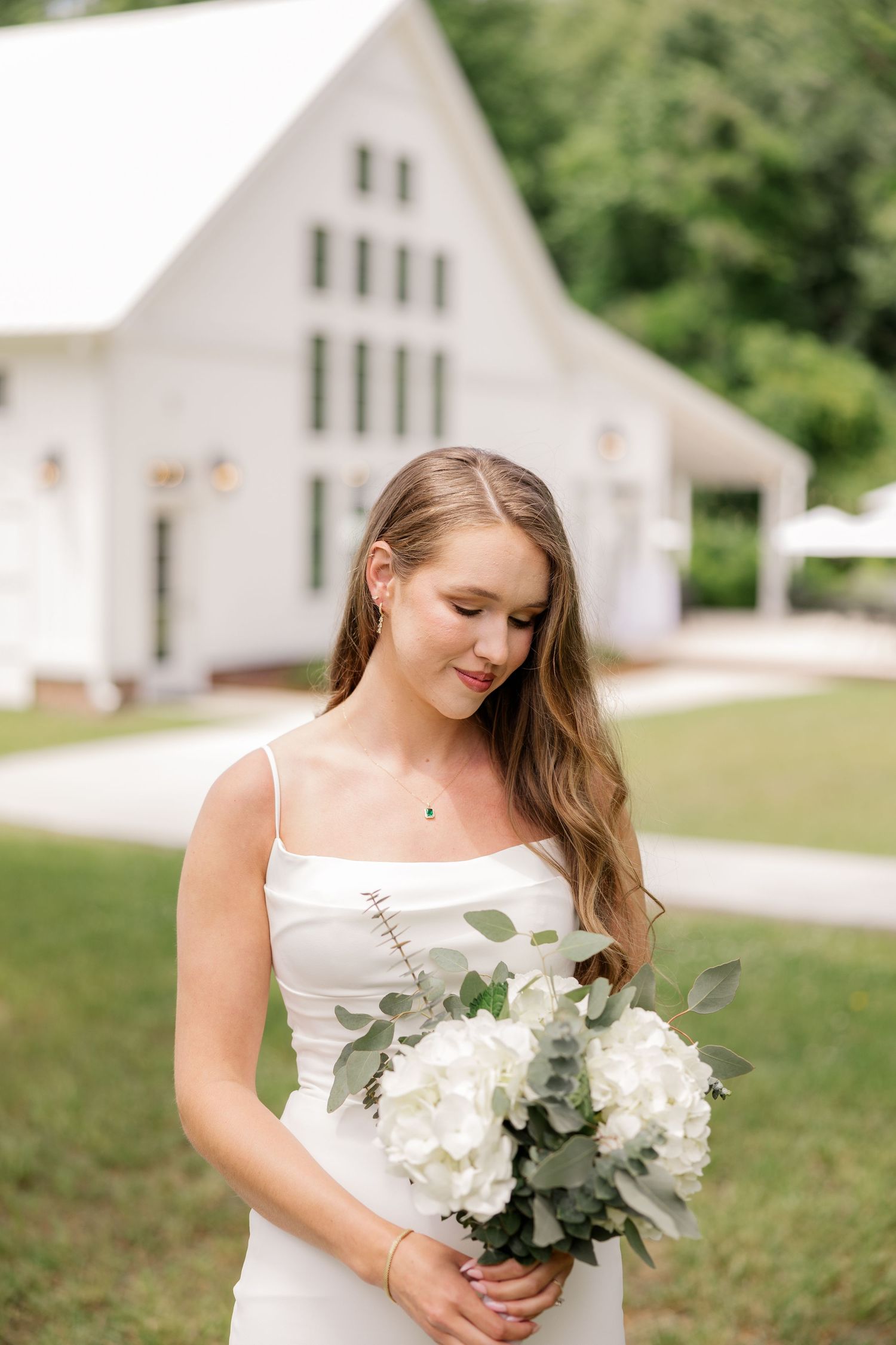 Bride in simple white dress holds elegant bouquet of white hydrangeas and eucalyptus with chapel in background.