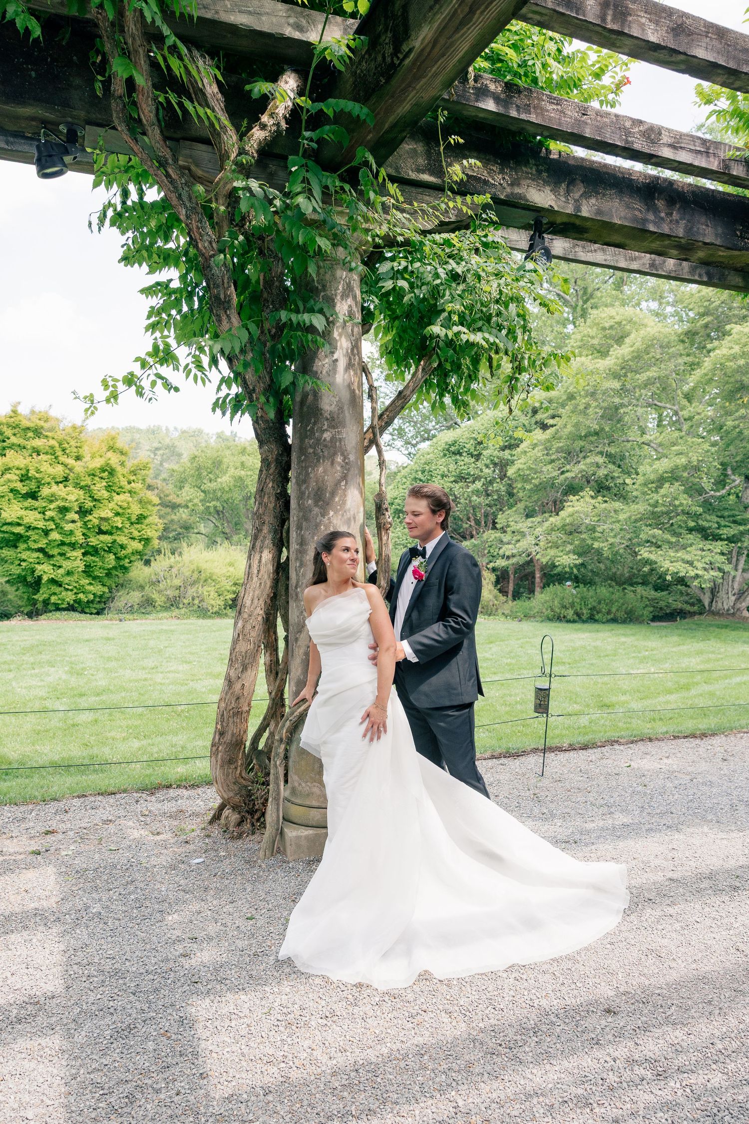 Bride and groom pose under a wooden pergola with lush greenery at an outdoor wedding venue.