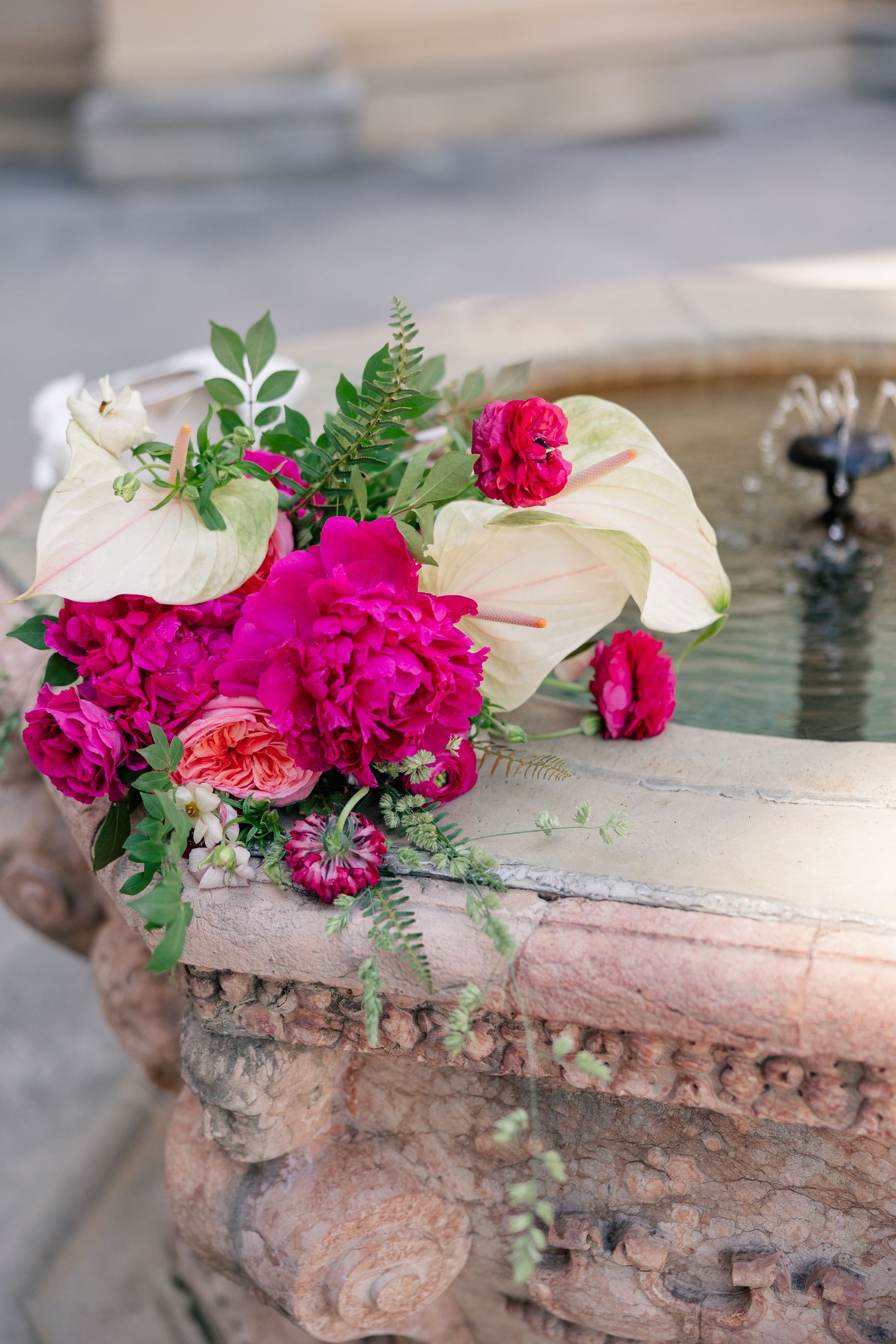 Vibrant pink peony and anthurium bridal bouquet resting on rustic stone fountain outdoors.