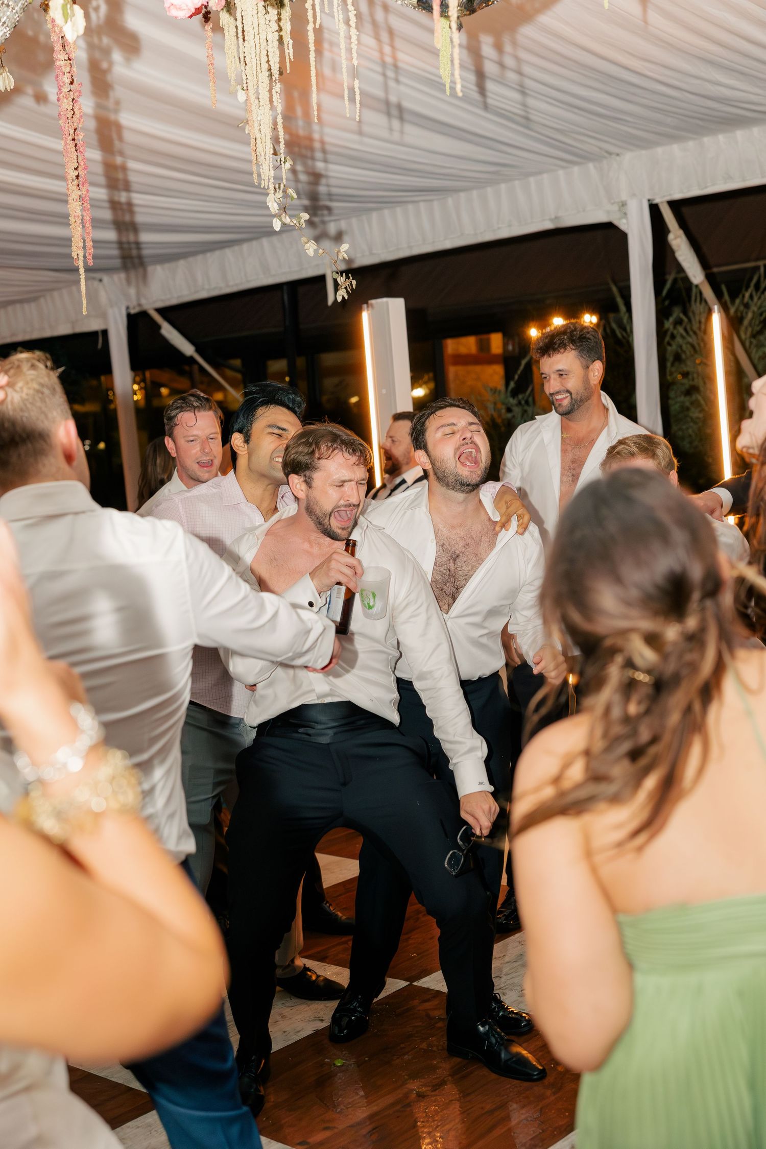 Groomsmen dancing energetically at a wedding reception under a white tent with elegant decor.
