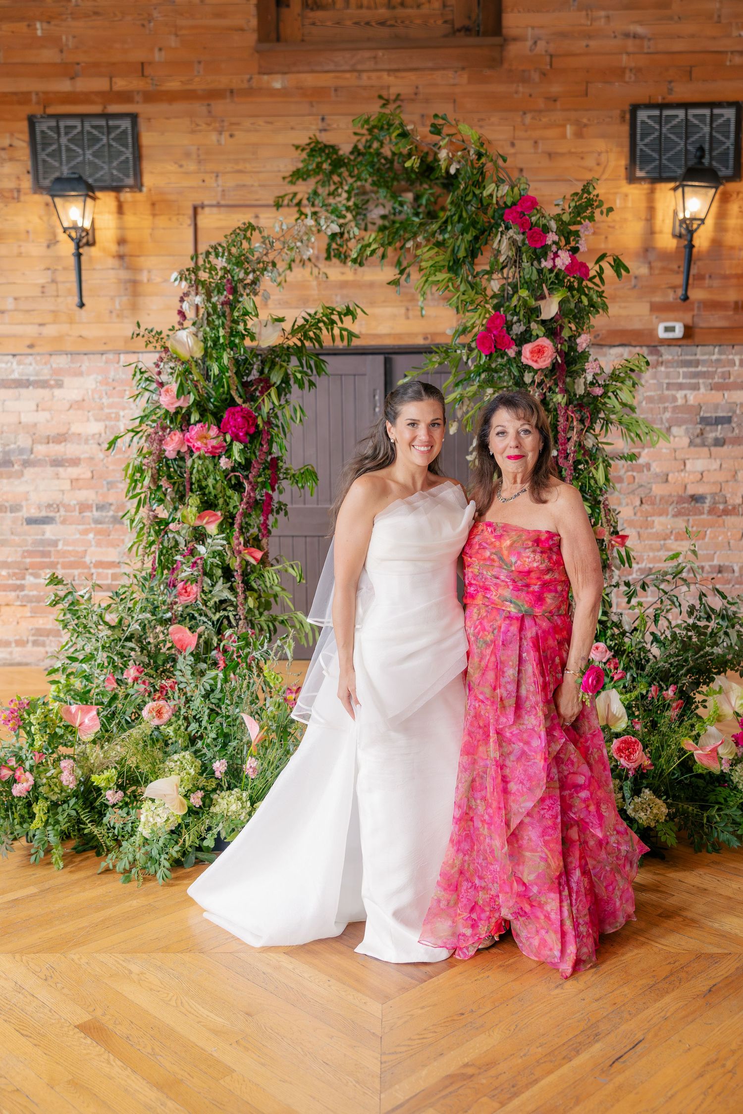 Bride and guest pose together in front of a stunning pink and green floral ceremony arch.