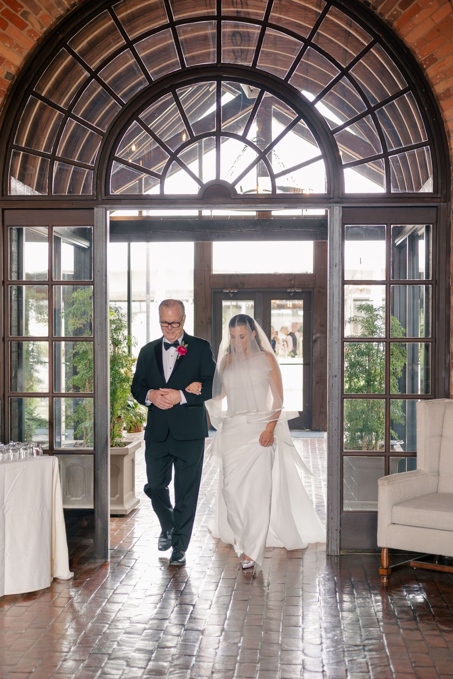 Bride walks down the aisle with her father through elegant arched glass doors at a wedding venue.