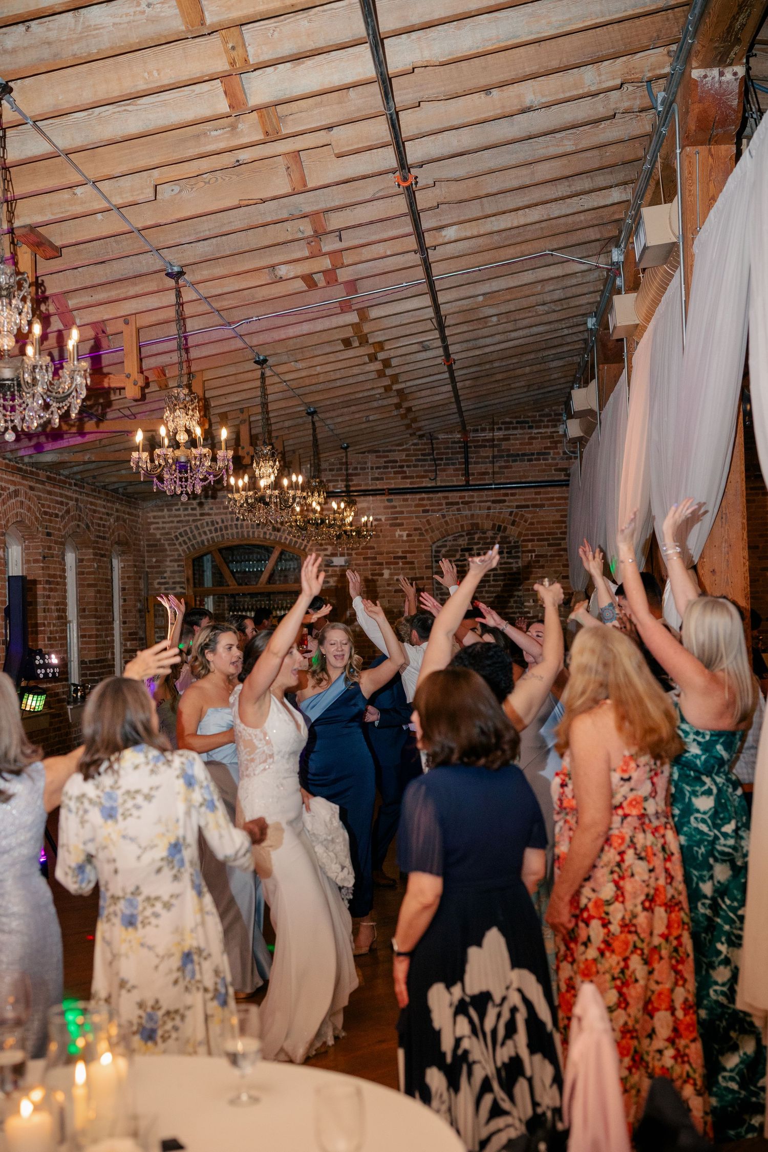 Wedding guests raise hands on dance floor at rustic brick venue with chandeliers.