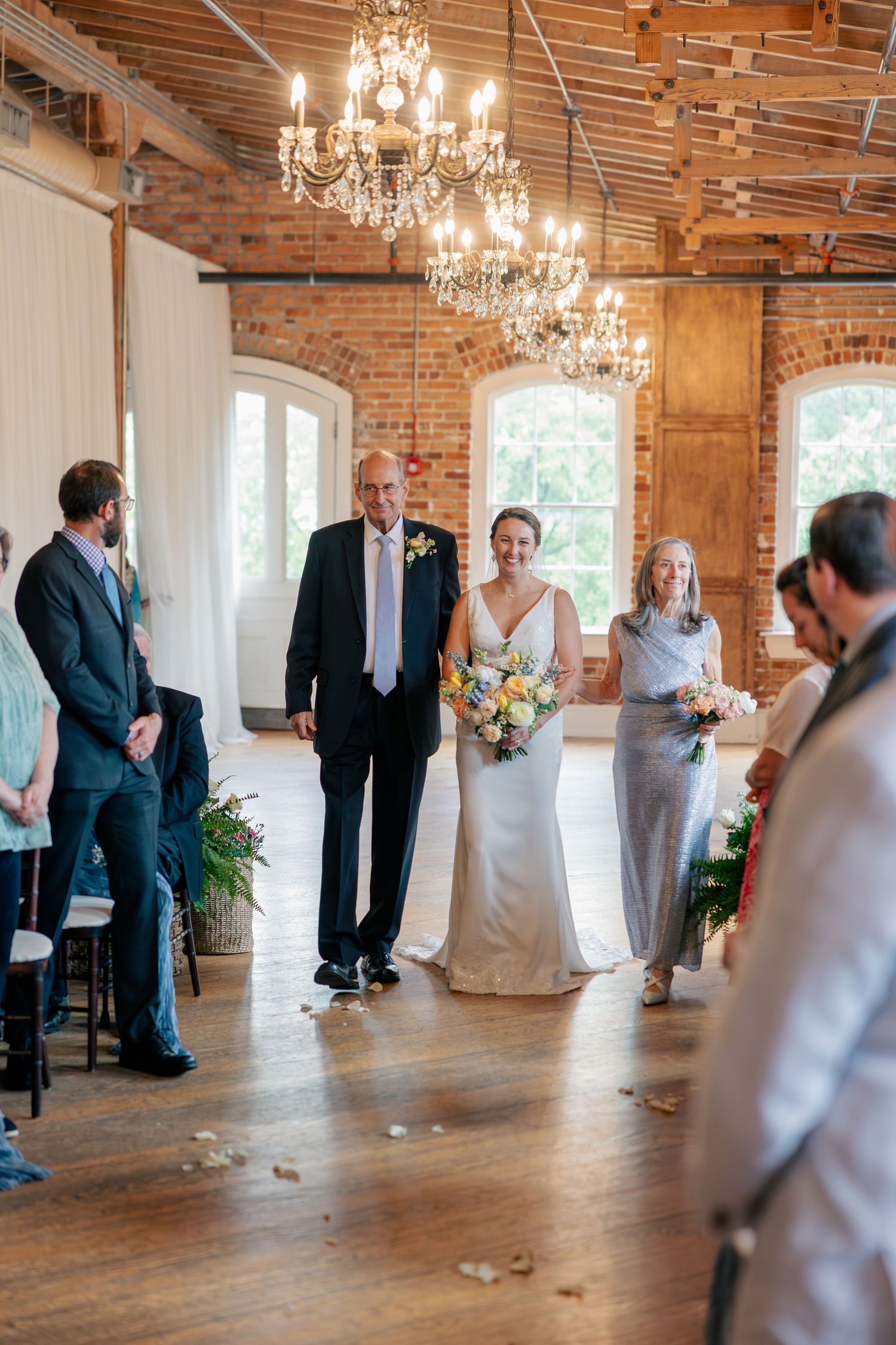 Radiant bride walks down aisle with parents in chandelier-lit rustic brick wedding venue.