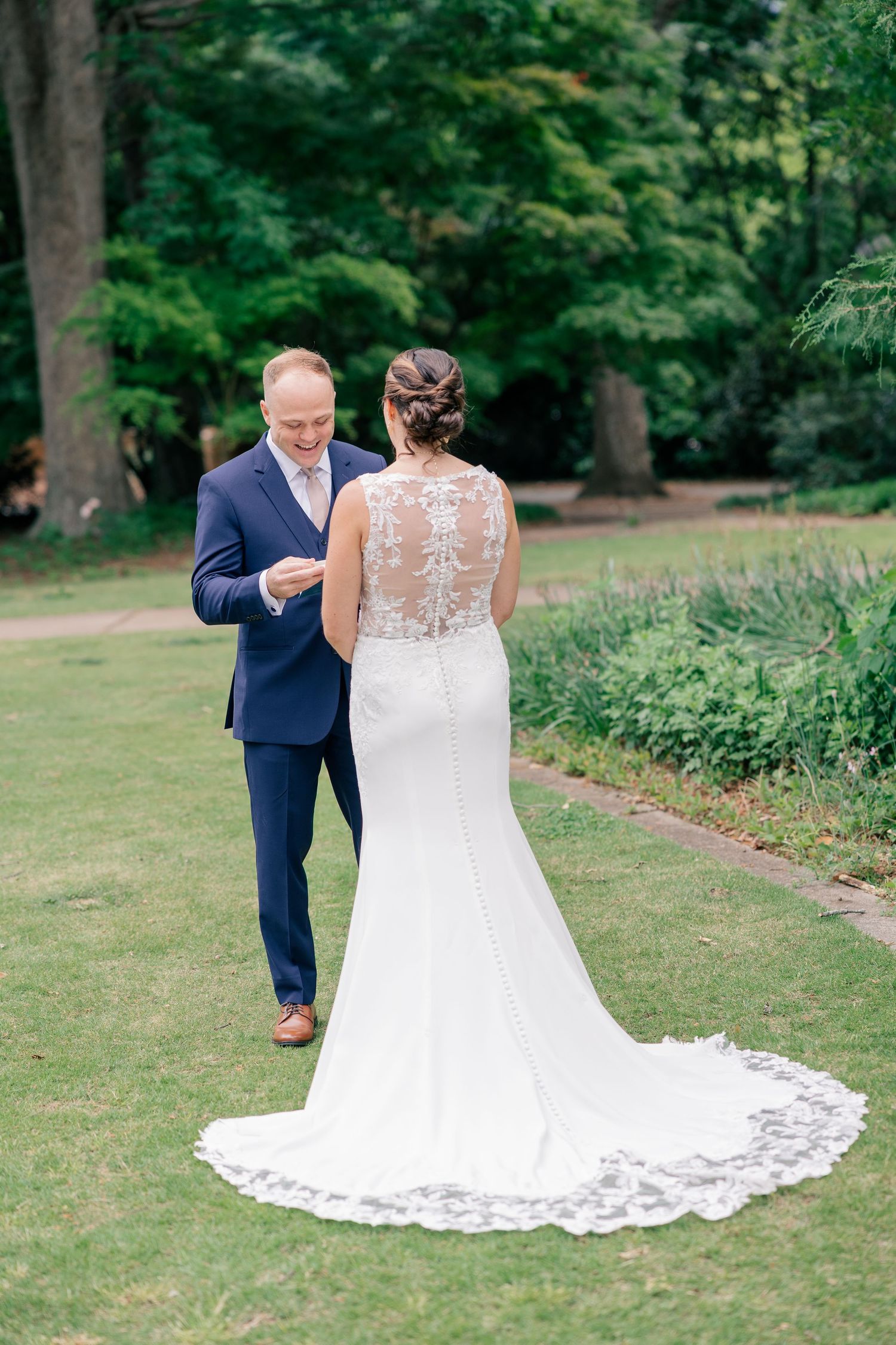 Bride in lace-back wedding gown shares first look moment with groom in navy suit in lush garden.