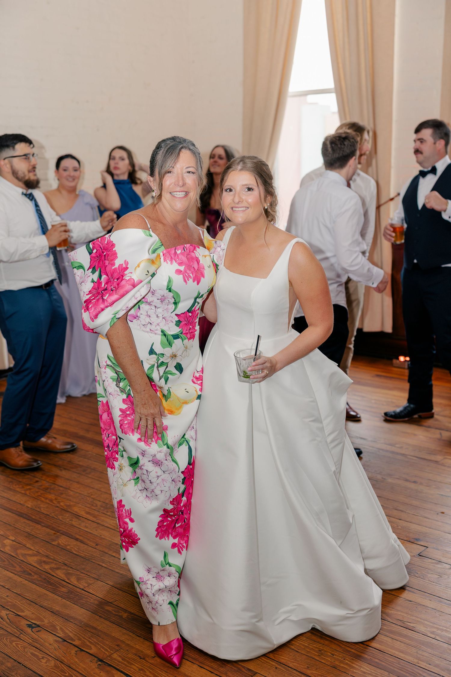 Bride in white gown poses with guest in floral dress at wedding reception on hardwood dance floor.