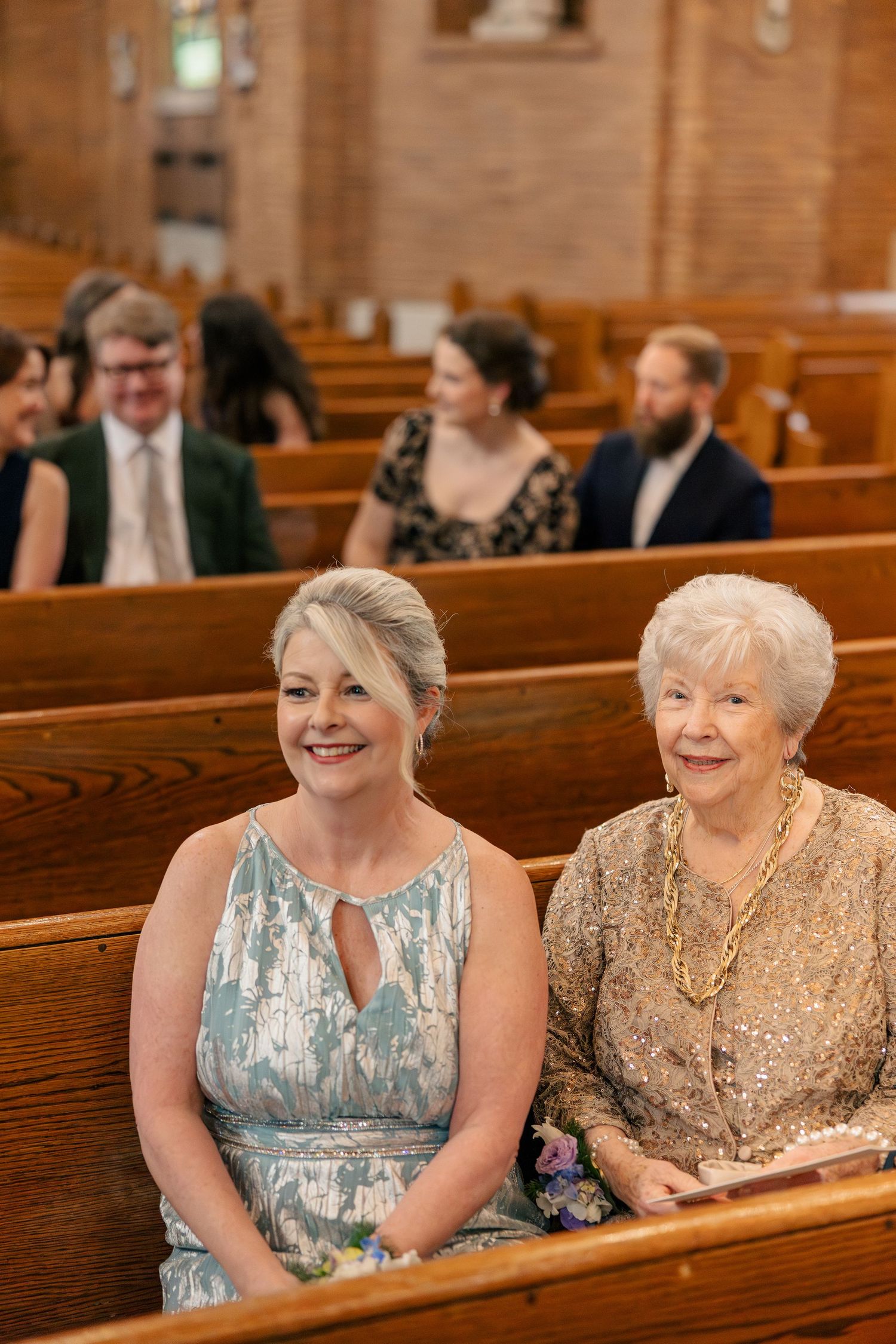 Two elegantly dressed women smile while seated in wooden church pews during a wedding ceremony.