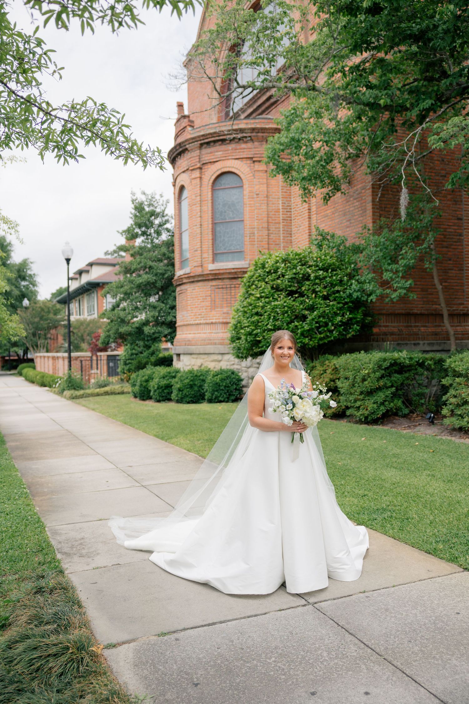 Bride in full white ball gown with cathedral veil holds bouquet outside historic red brick church.
