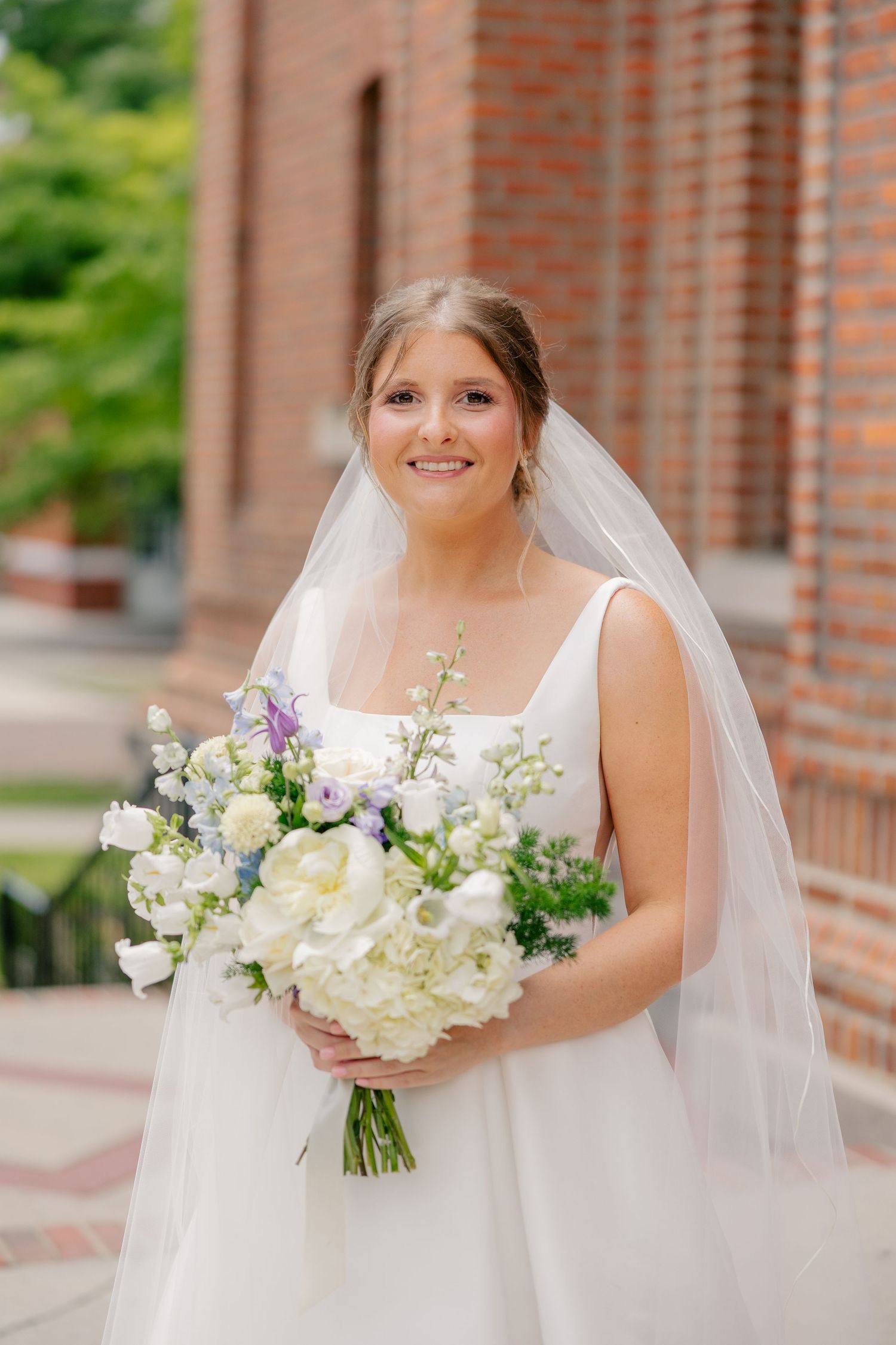 Smiling bride in white square-neck gown and veil holds bouquet of white and lavender flowers outside brick church.
