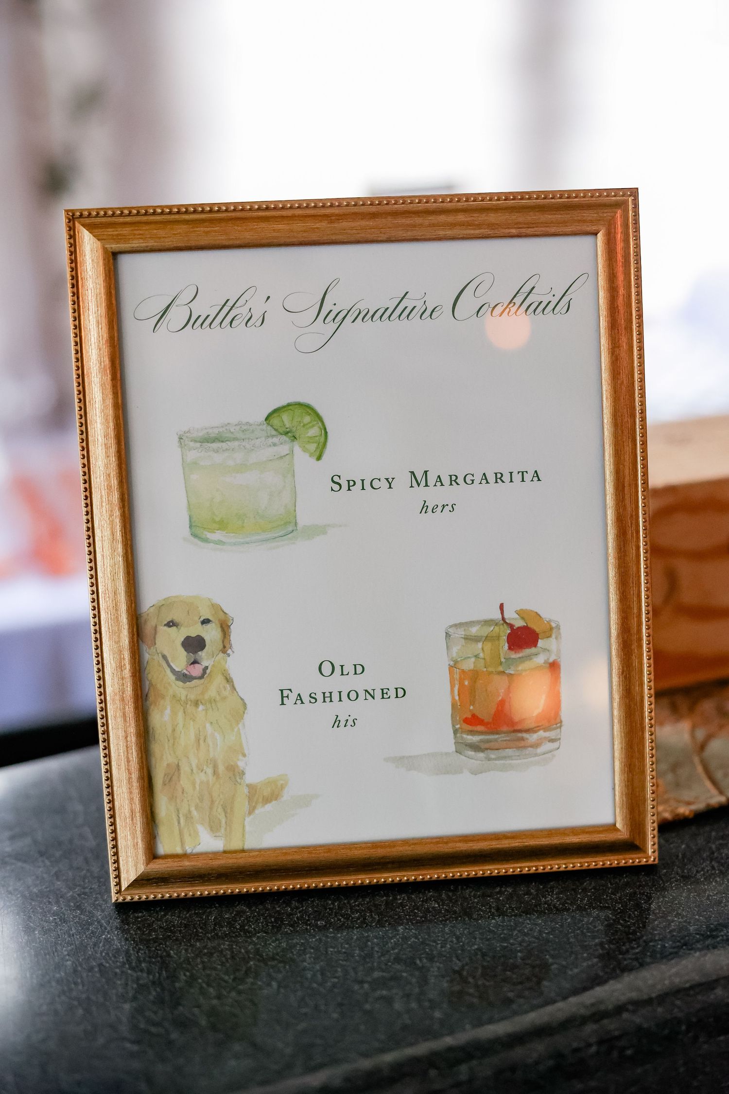 Gold-framed sign displaying Butler's Signature Cocktails menu featuring a Spicy Margarita and Old Fashioned with watercolor illustrations.