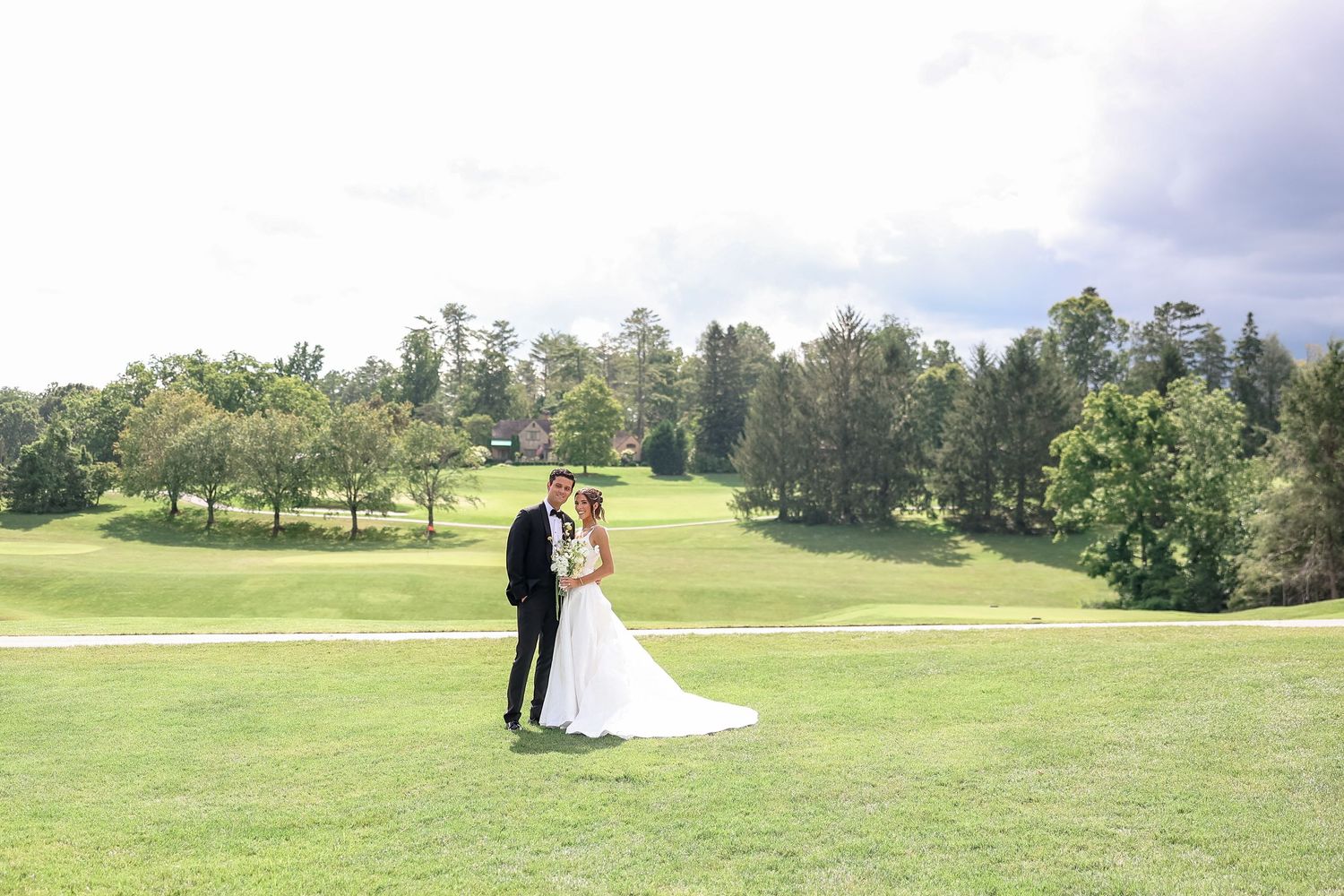Bride and groom pose together on scenic golf course lawn surrounded by lush trees on their wedding day.