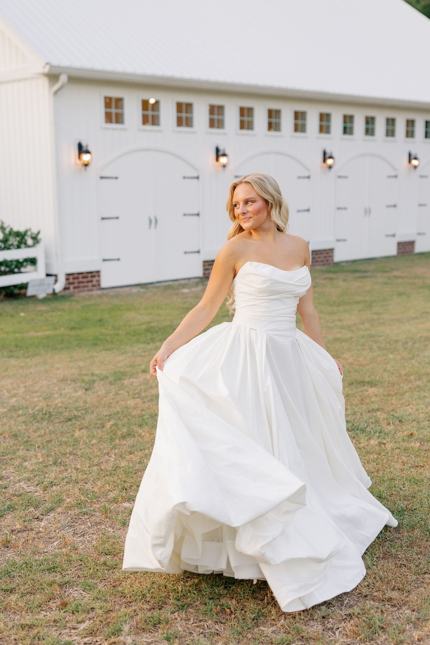 Bride twirling in strapless white ballgown outside white barn venue in golden hour light.
