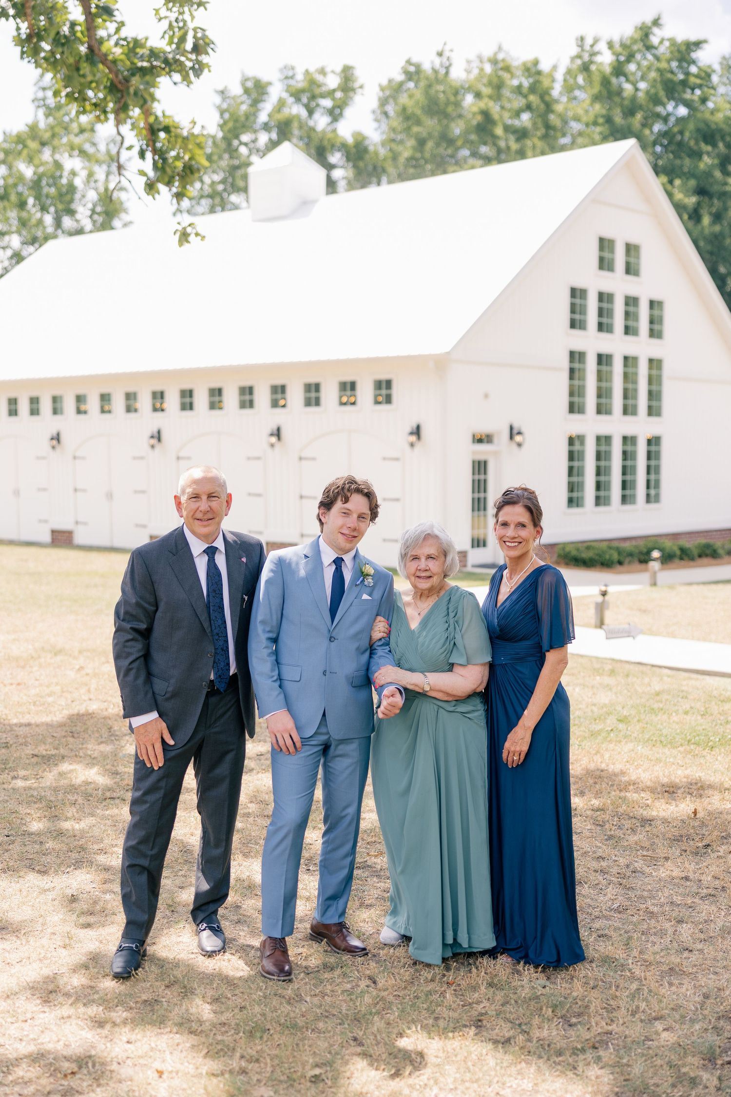 Groom in light blue suit poses with three family members in front of a white barn wedding venue.