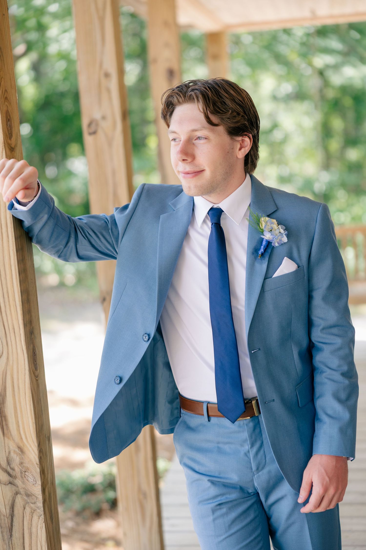 Groom in a light blue suit with navy tie and boutonniere leaning against a wooden post outdoors.