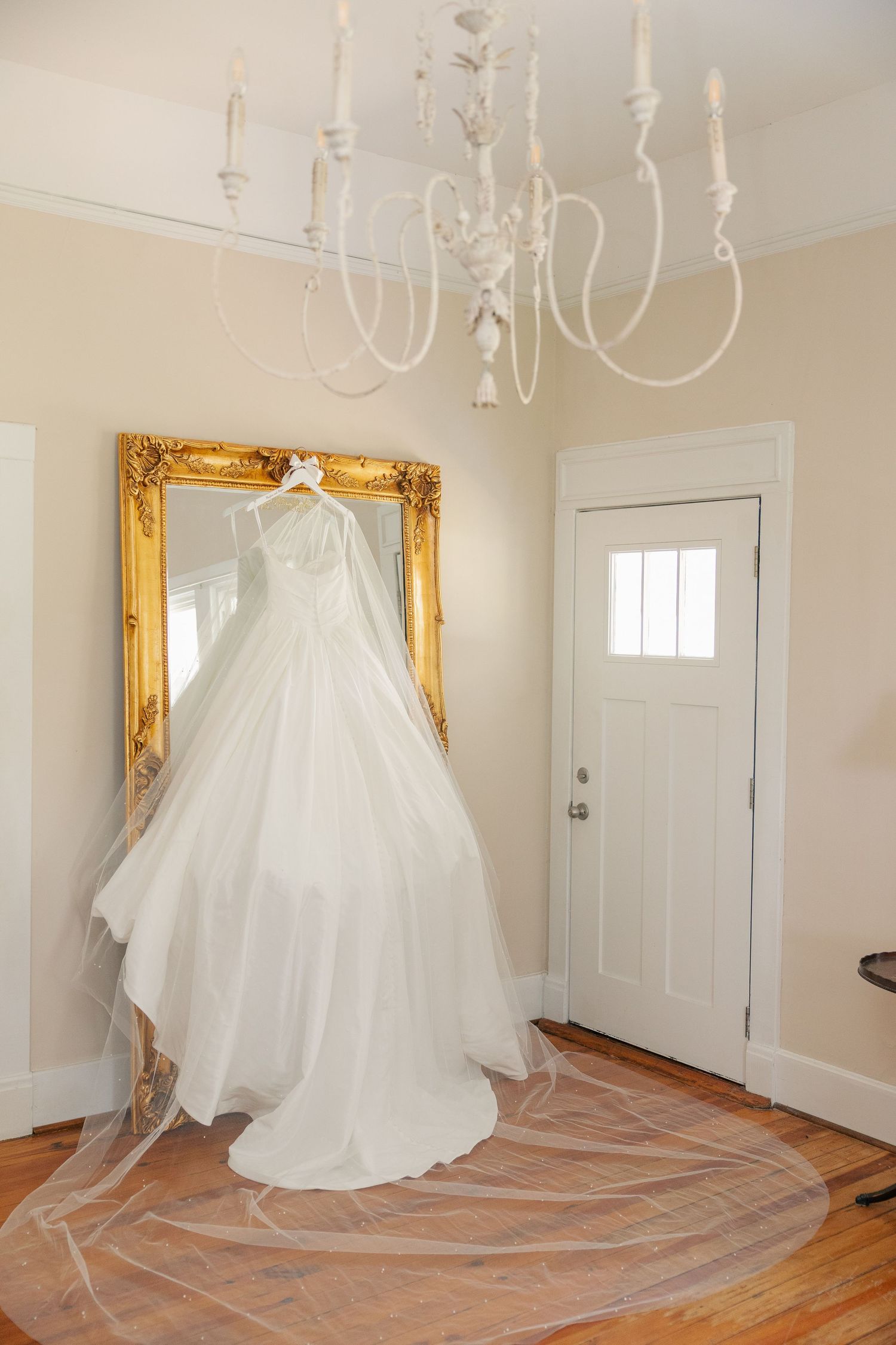 White wedding gown with long veil hanging on gold ornate mirror in elegant entryway with chandelier.