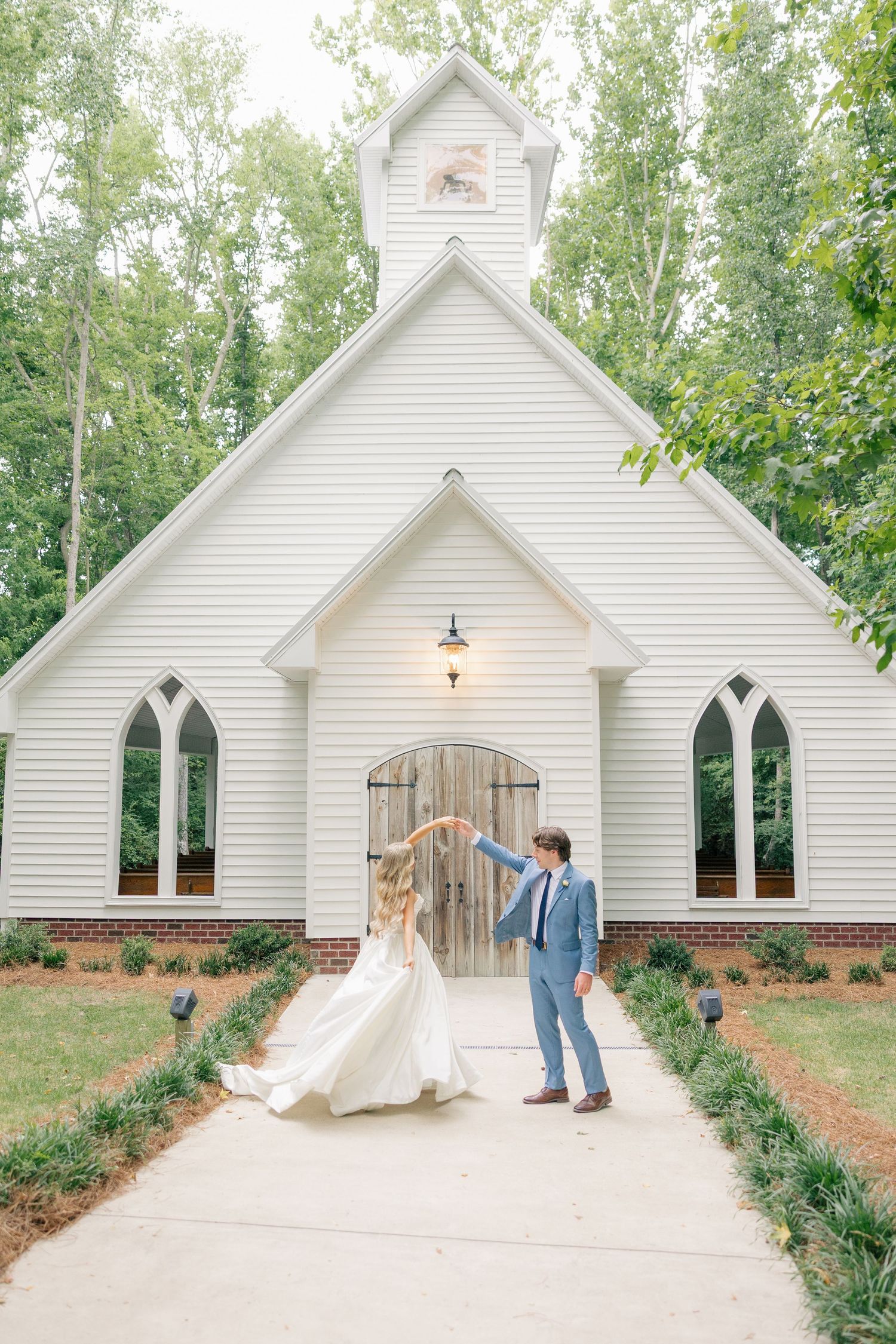 Bride and groom dance together on pathway in front of charming white chapel surrounded by trees.