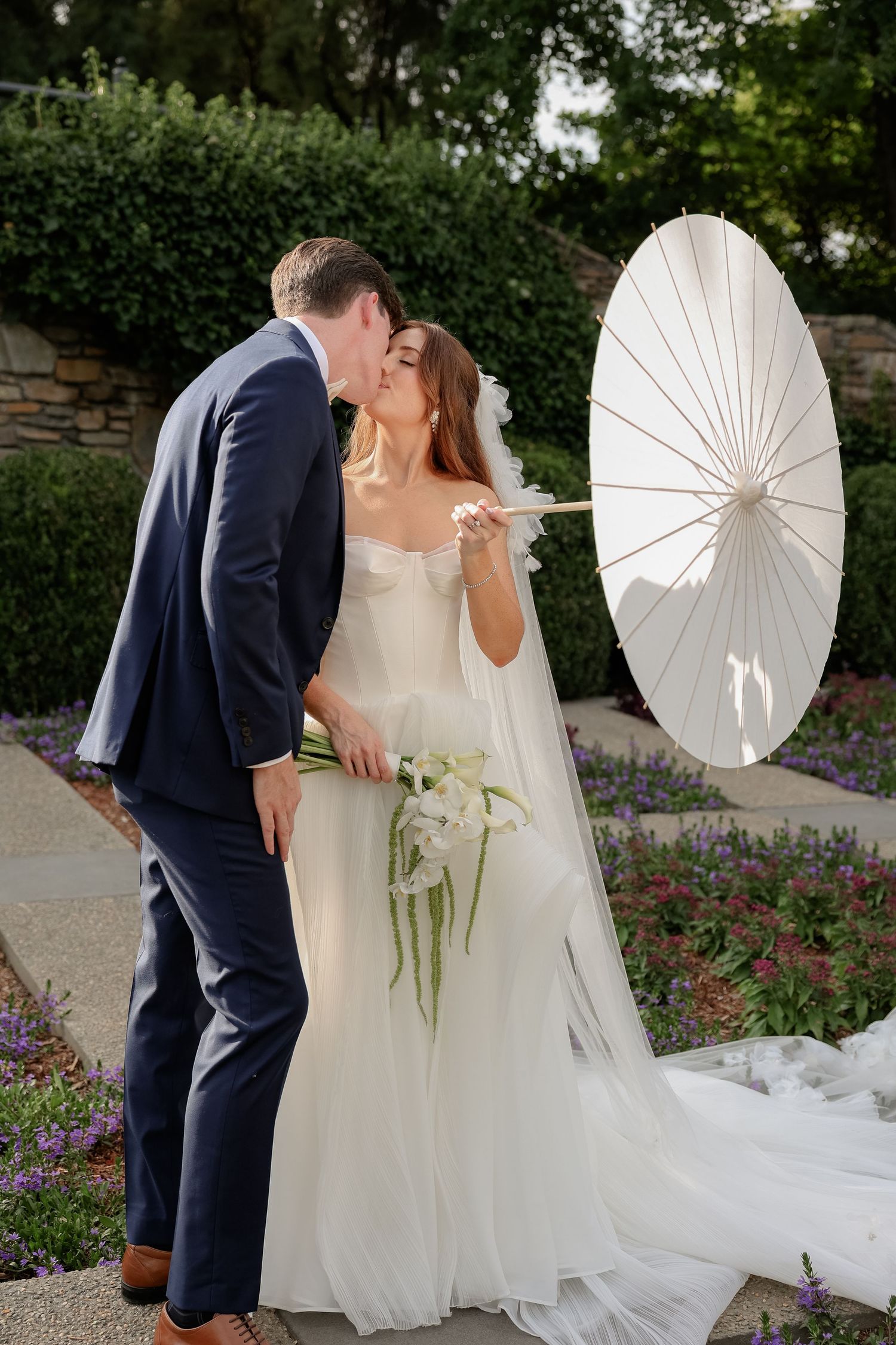 A romantic wedding photo session in a garden featuring a white parasol and flowing bridal gown.