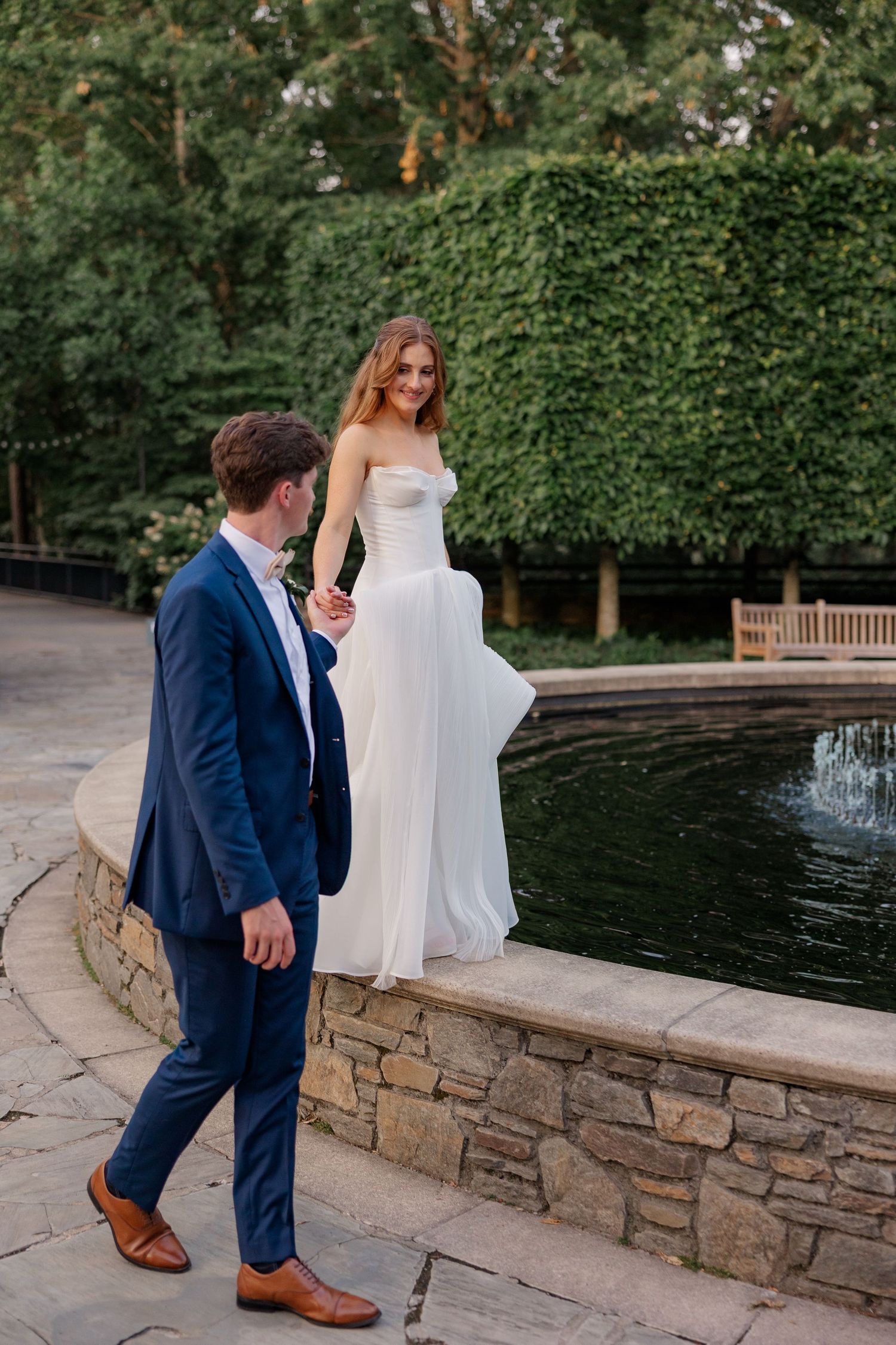 A couple in wedding attire stands by a stone wall fountain in a garden setting.