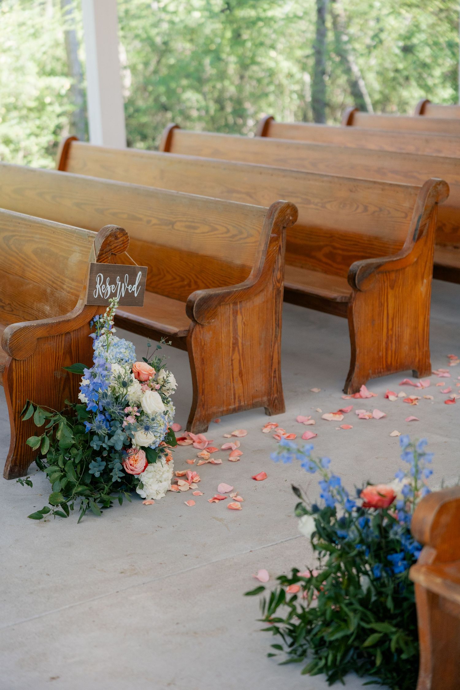 Wooden church pews decorated with pink and blue floral arrangements for a wedding ceremony.