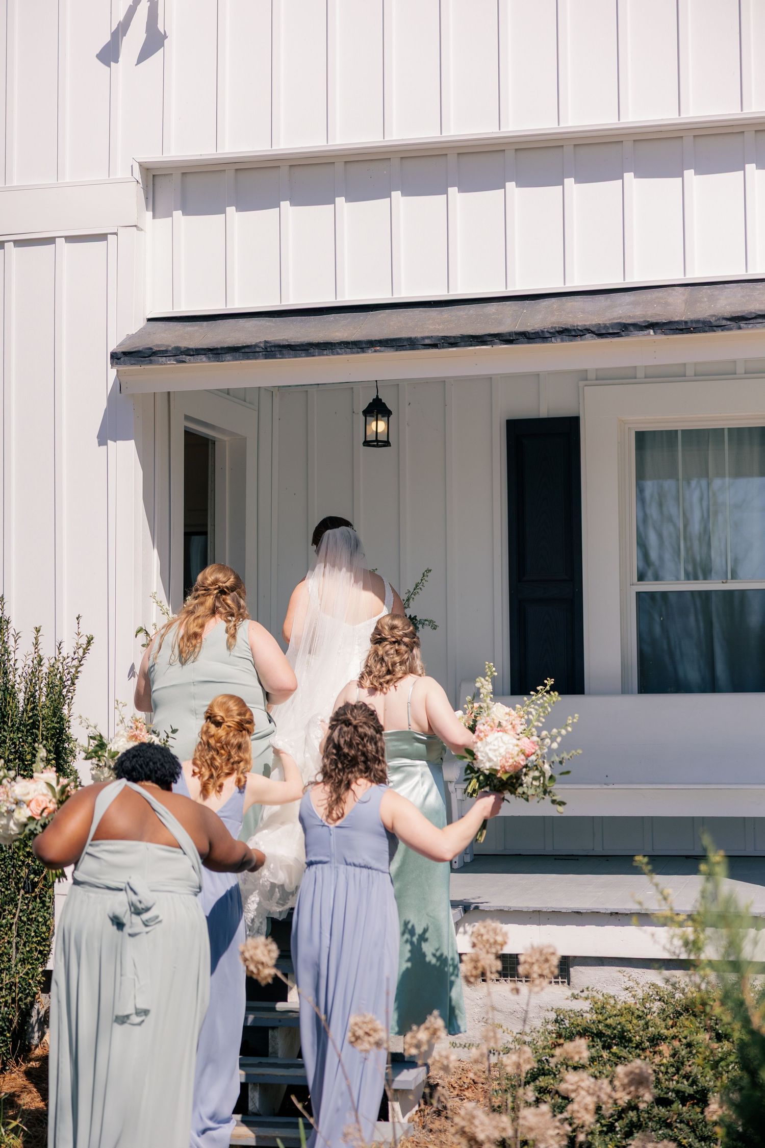 Bridesmaids in light blue dresses wave goodbye outside a white farmhouse-style building.
