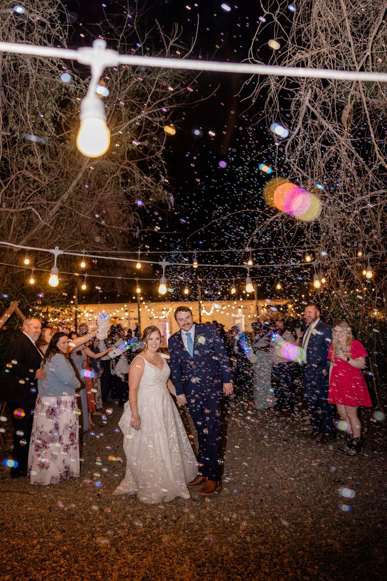 Newlyweds walk under string lights while guests shower them with confetti during an evening celebration.