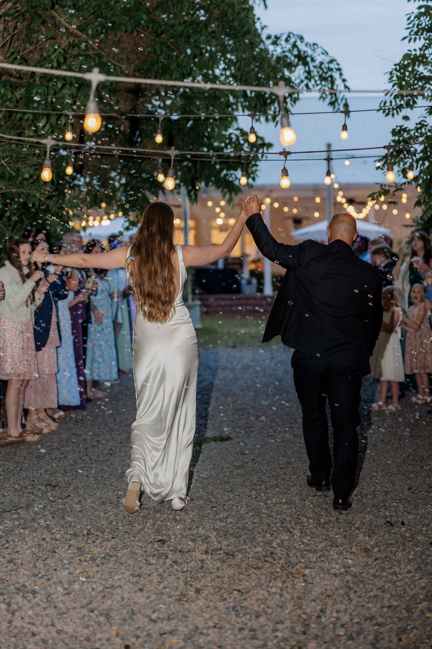 Wedding couple exits venue under twinkling lights while guests celebrate with confetti.