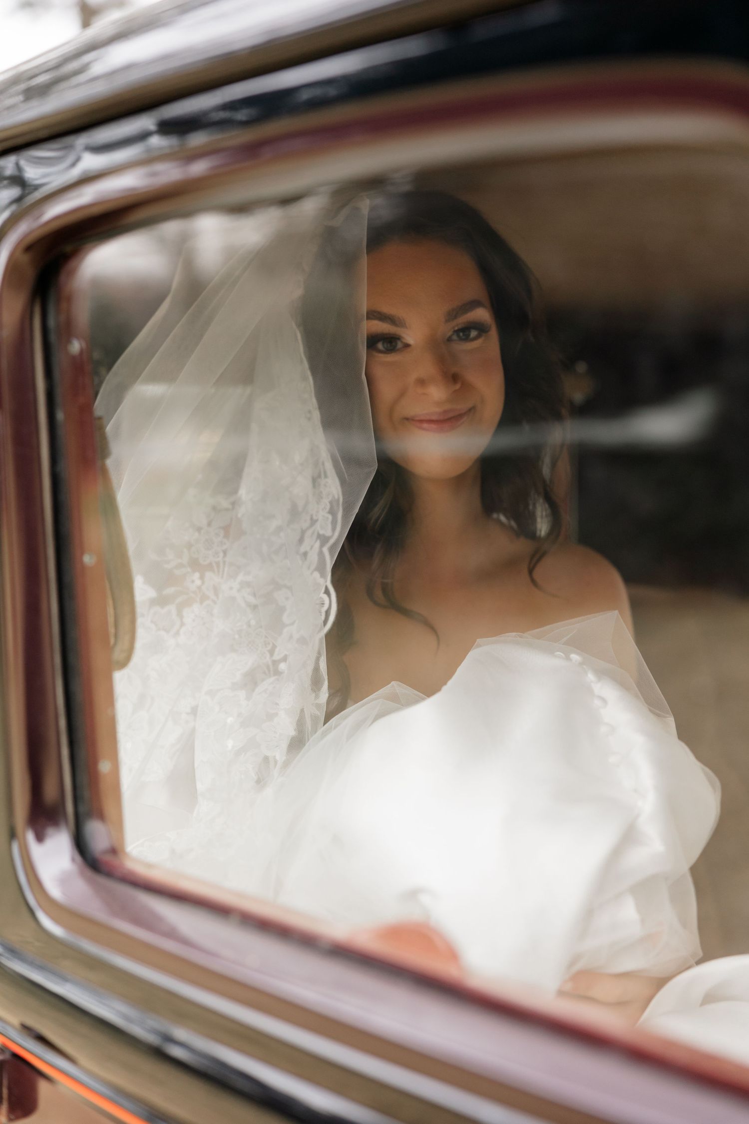 Bride in white dress seen through vintage car window with soft focus ethereal effect.