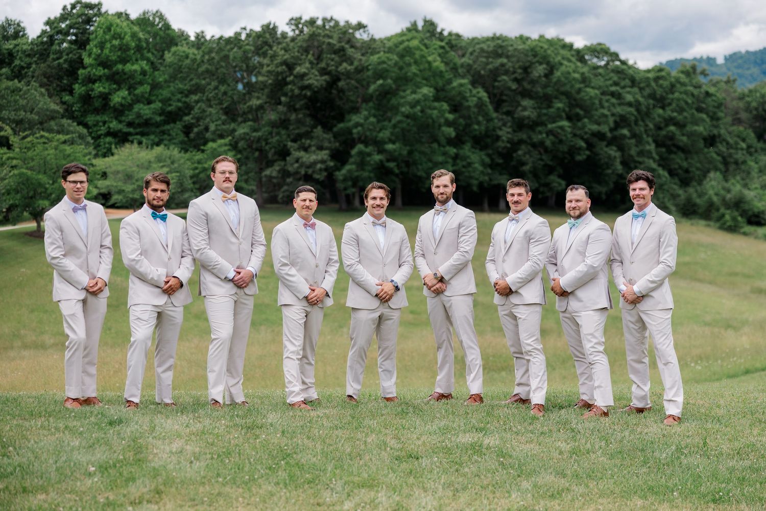 Wedding party members in light colored suits stand in a row on a grassy hillside for formal group photos.