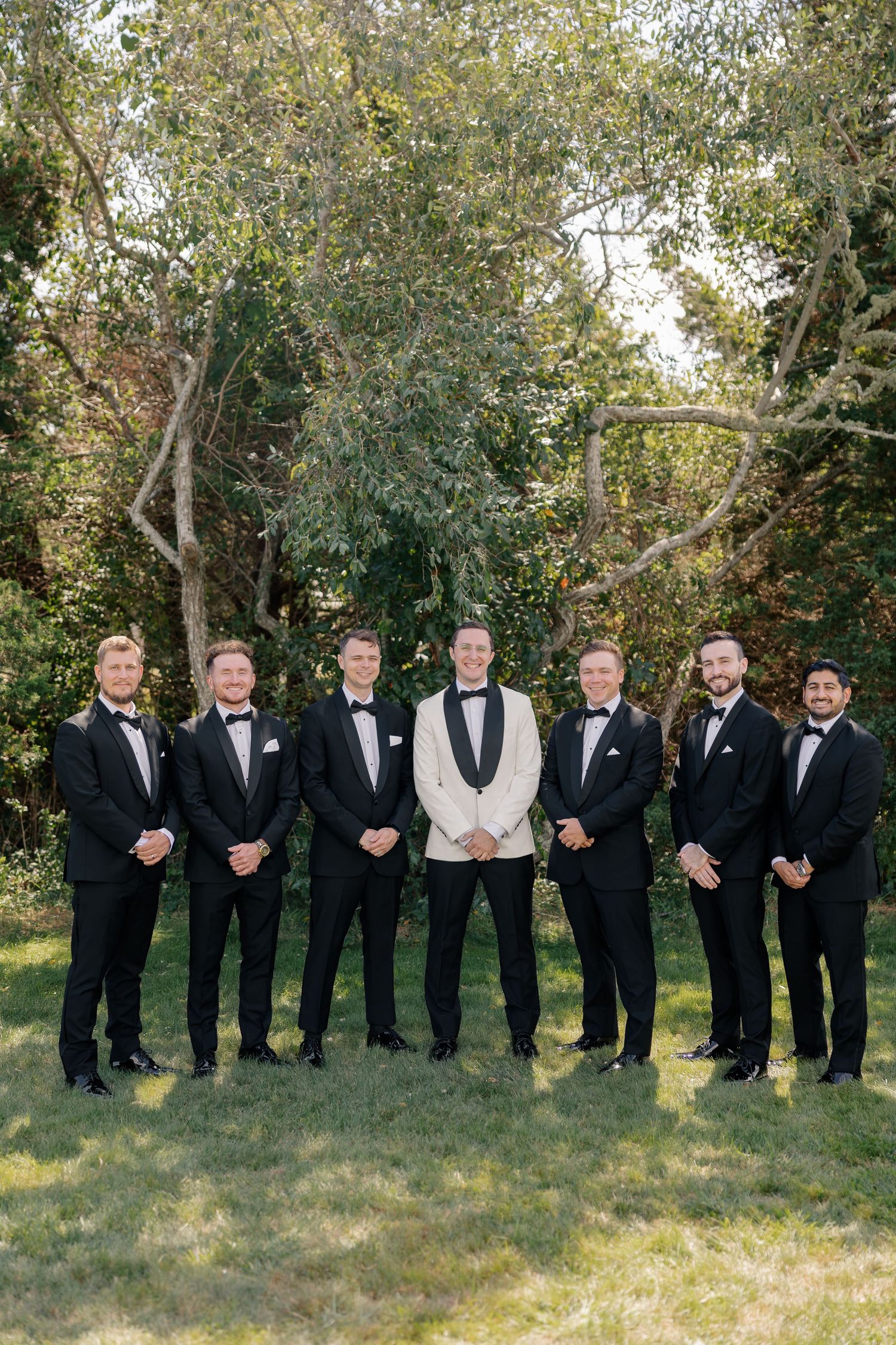 Groom in a white tuxedo jacket stands with six groomsmen in matching black tuxedos on a green lawn.