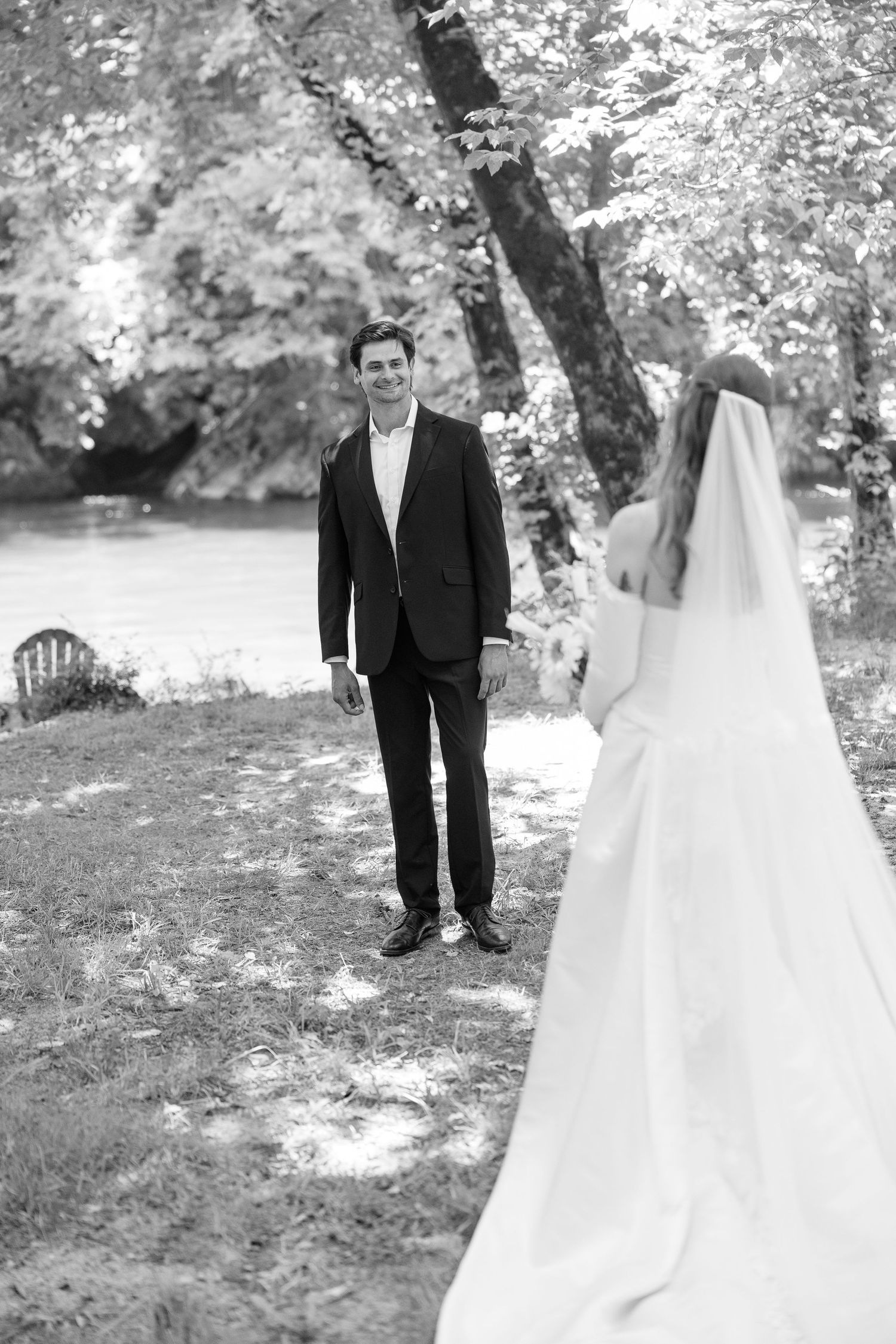 Black and white first look photo of smiling groom seeing bride by a riverside.