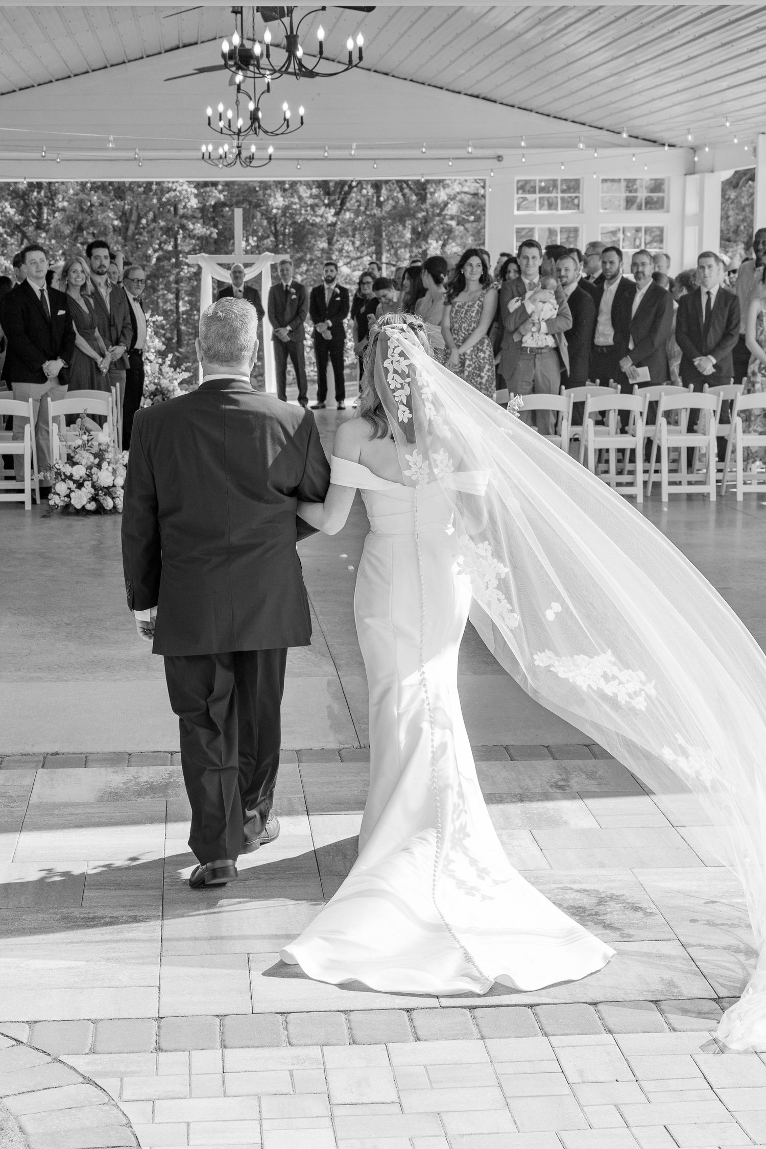 Black and white photo of bride walking down aisle with escort at outdoor wedding ceremony.