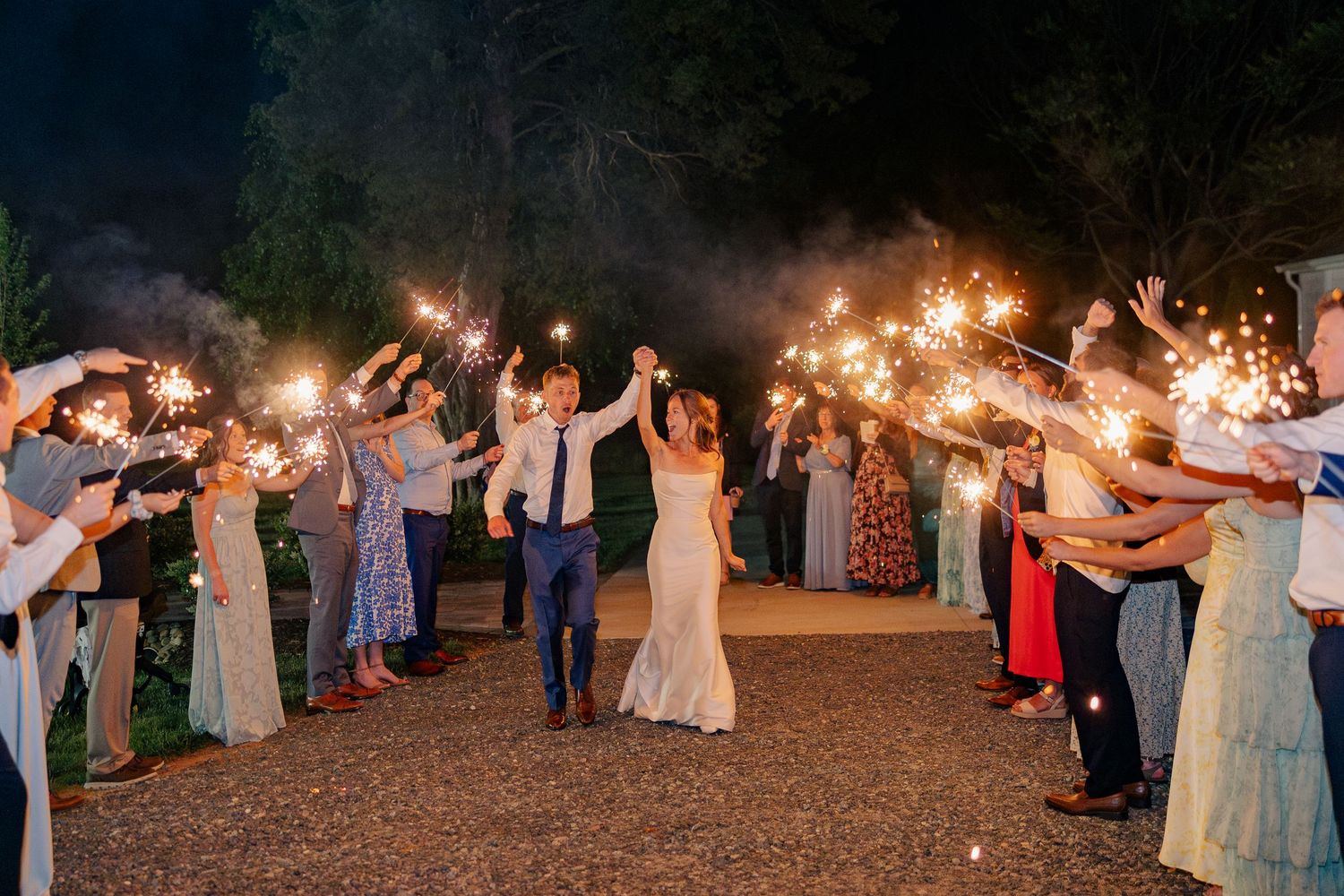 Bride and groom exit through sparkler tunnel surrounded by cheering guests at nighttime wedding celebration.