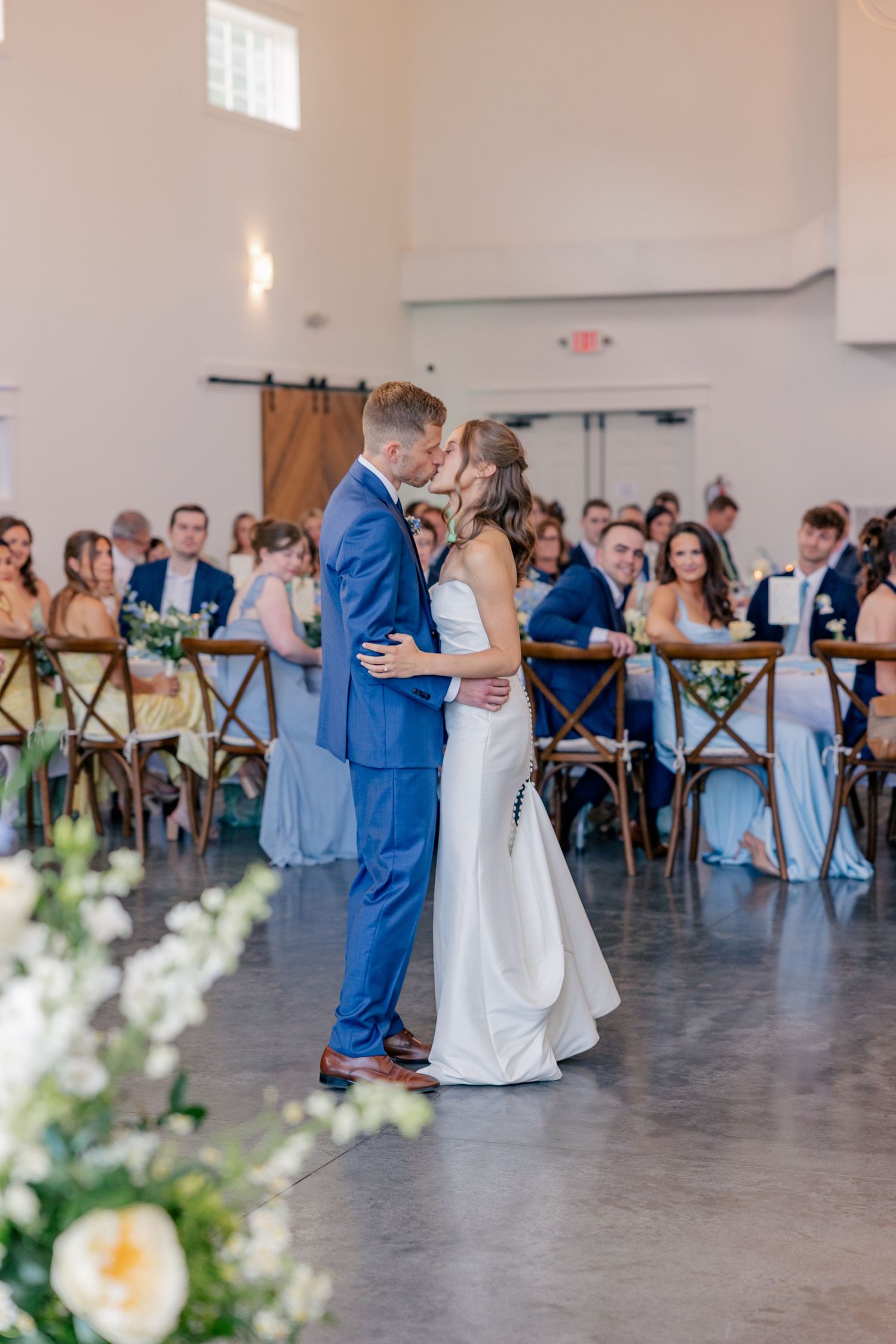 Bride and groom share first dance kiss in bright white barn reception hall with seated guests watching.