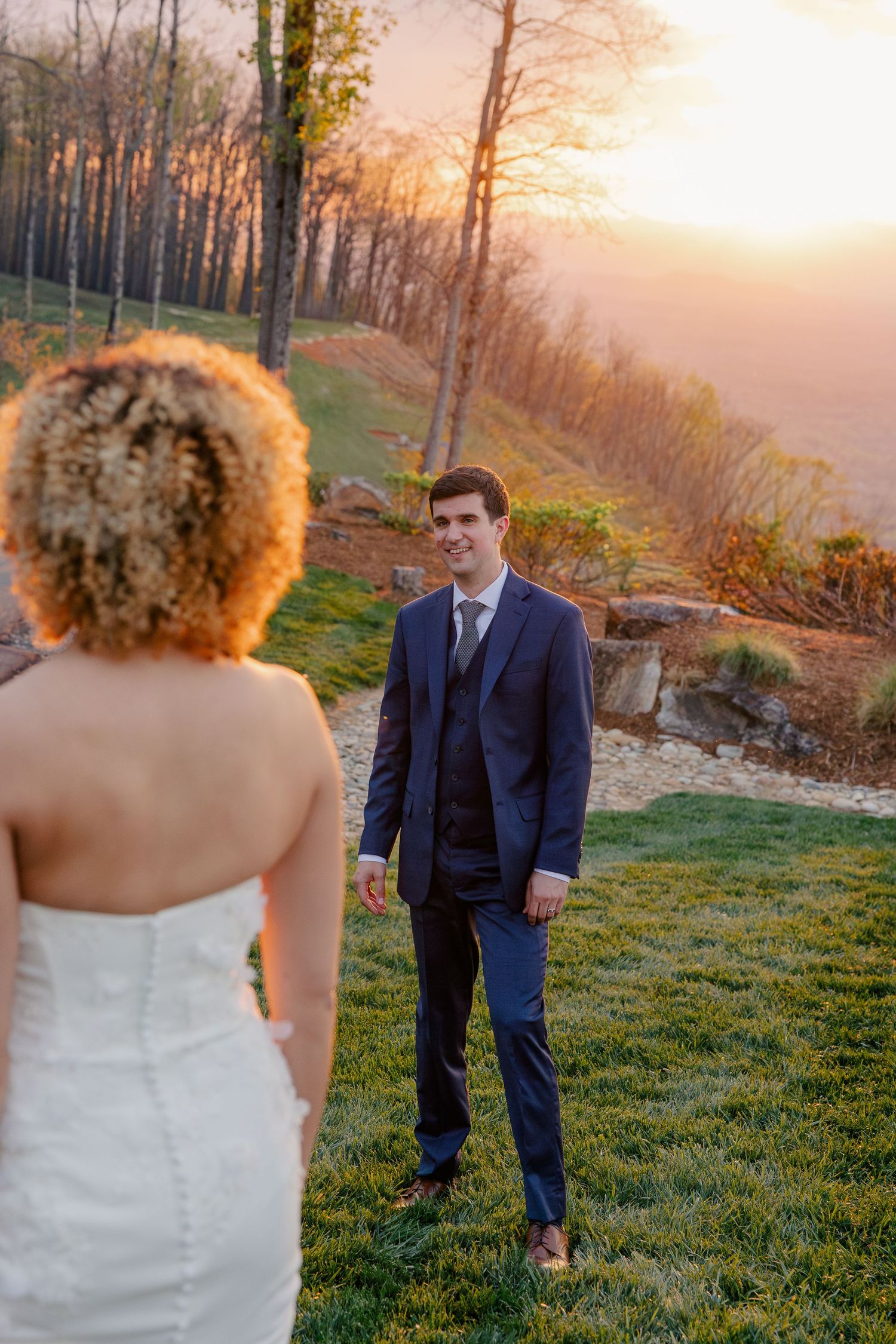 Groom in navy suit smiles at bride during golden sunset portraits with scenic mountain landscape backdrop.