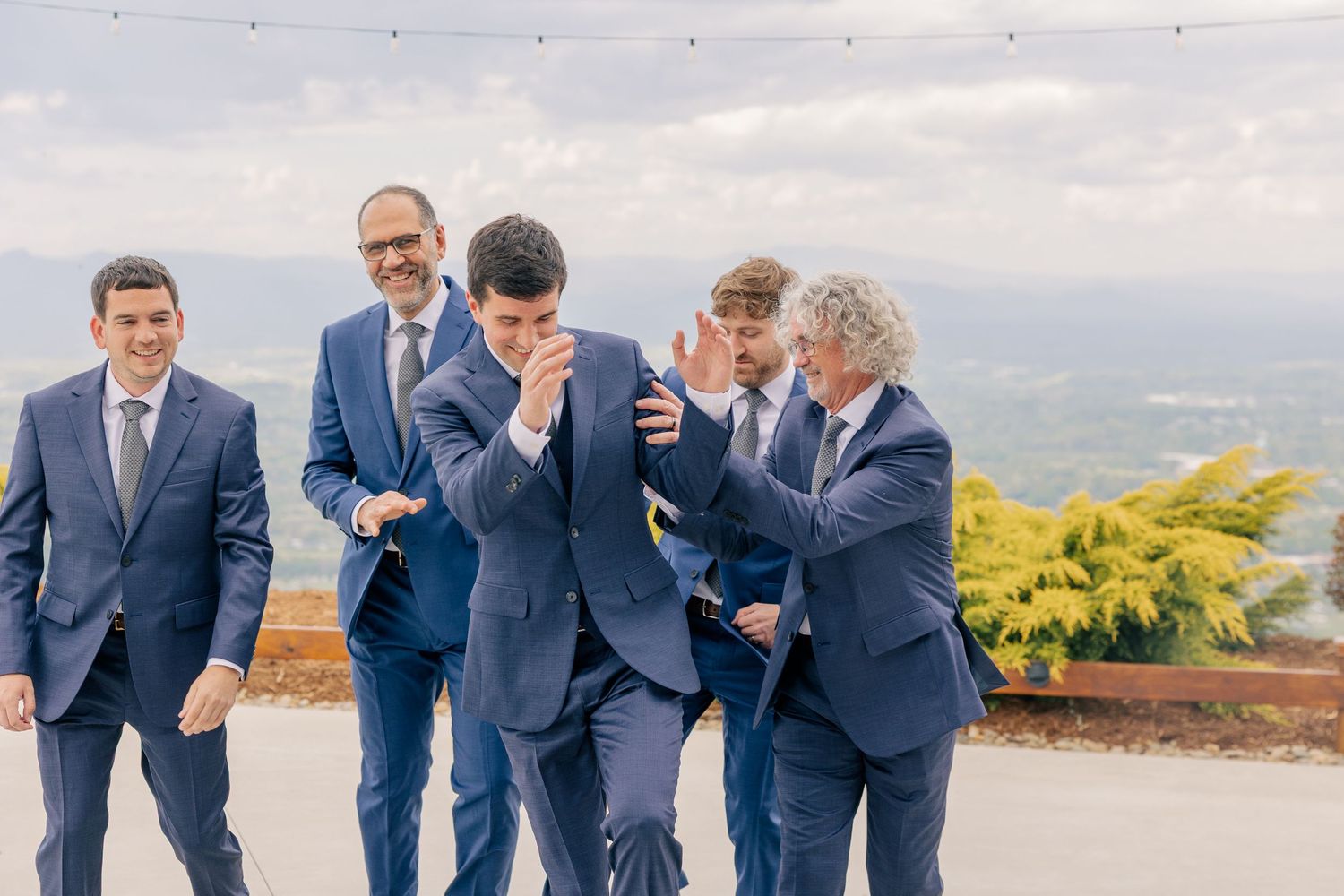 Groom laughing with groomsmen in navy suits on an outdoor terrace with sweeping mountain views.