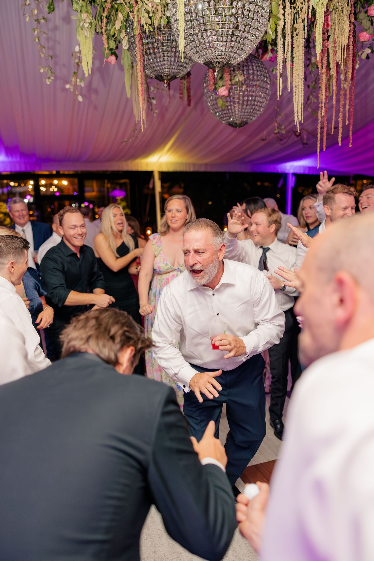 Energetic wedding guest dances joyfully on crowded dance floor under chandelier at evening reception.