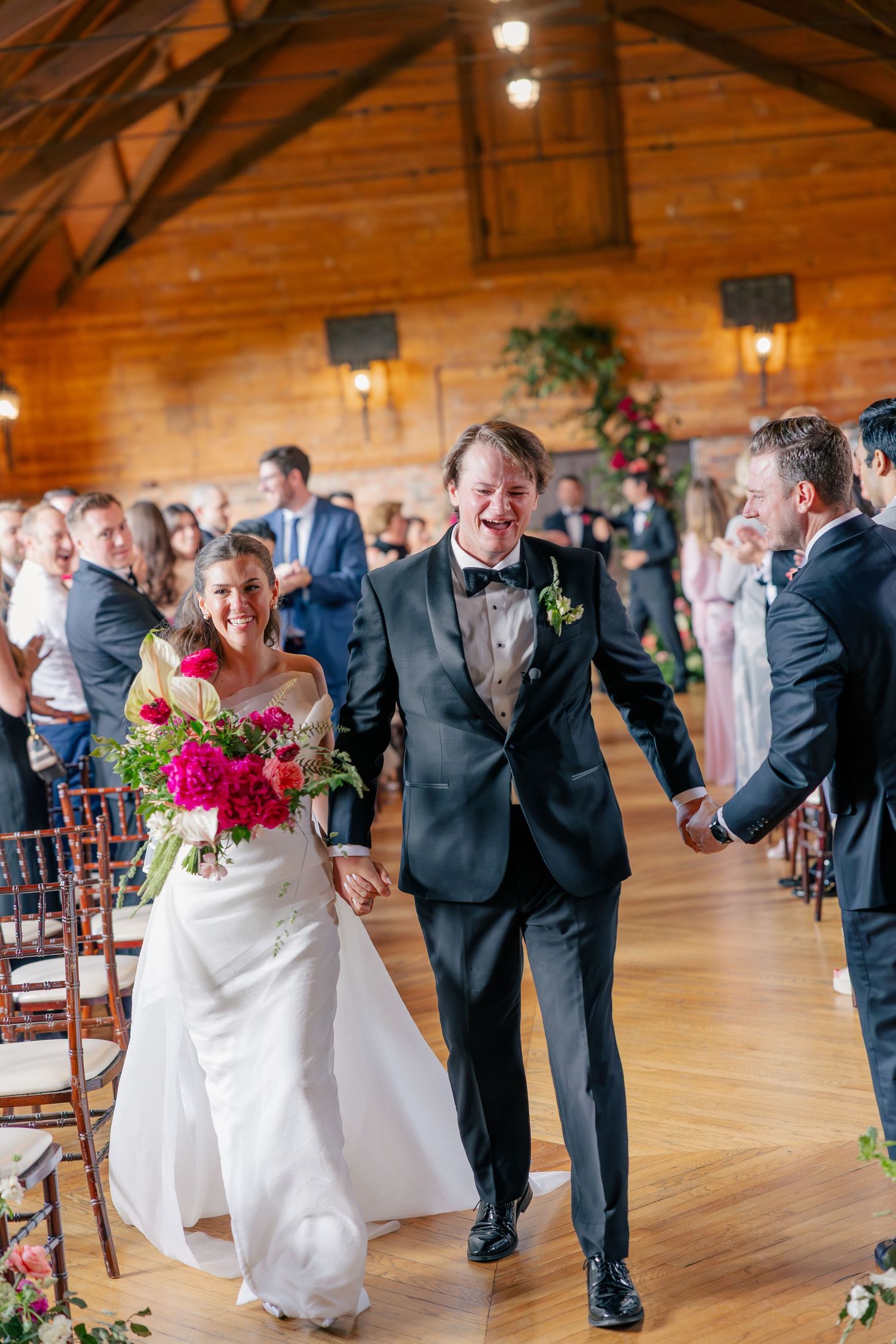 Laughing bride and groom walk down aisle after ceremony in rustic wooden wedding venue.