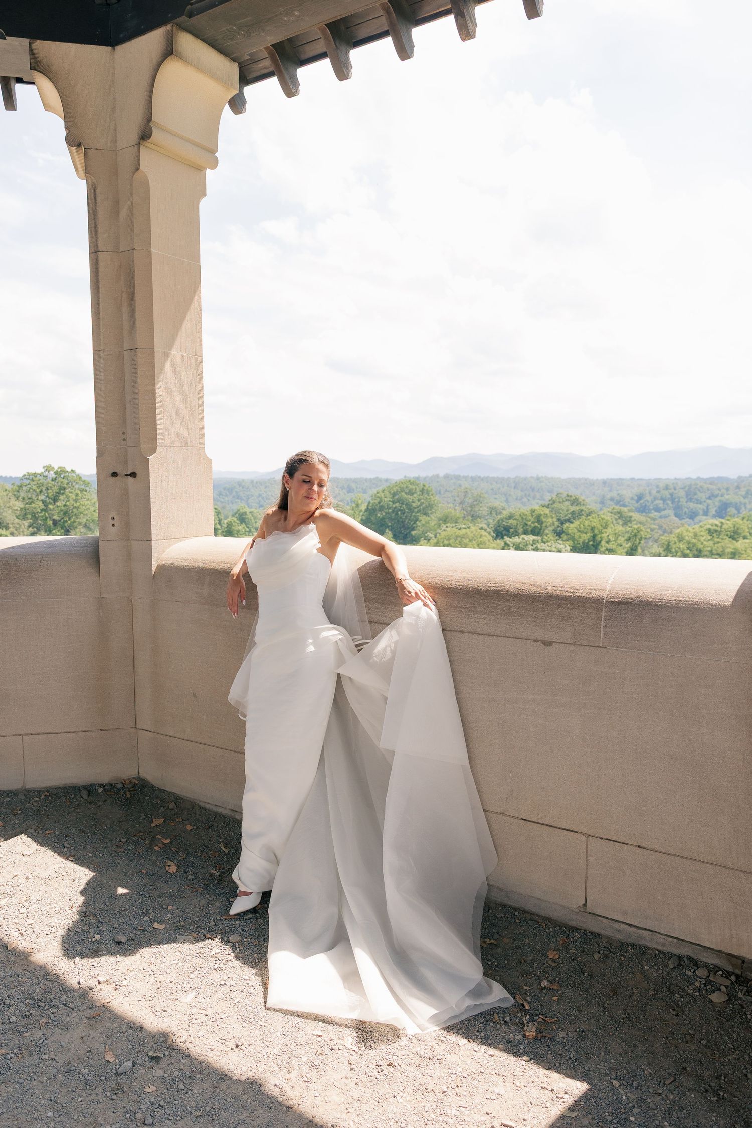 Bride in white gown with flowing train poses on stone balcony overlooking lush green mountains.