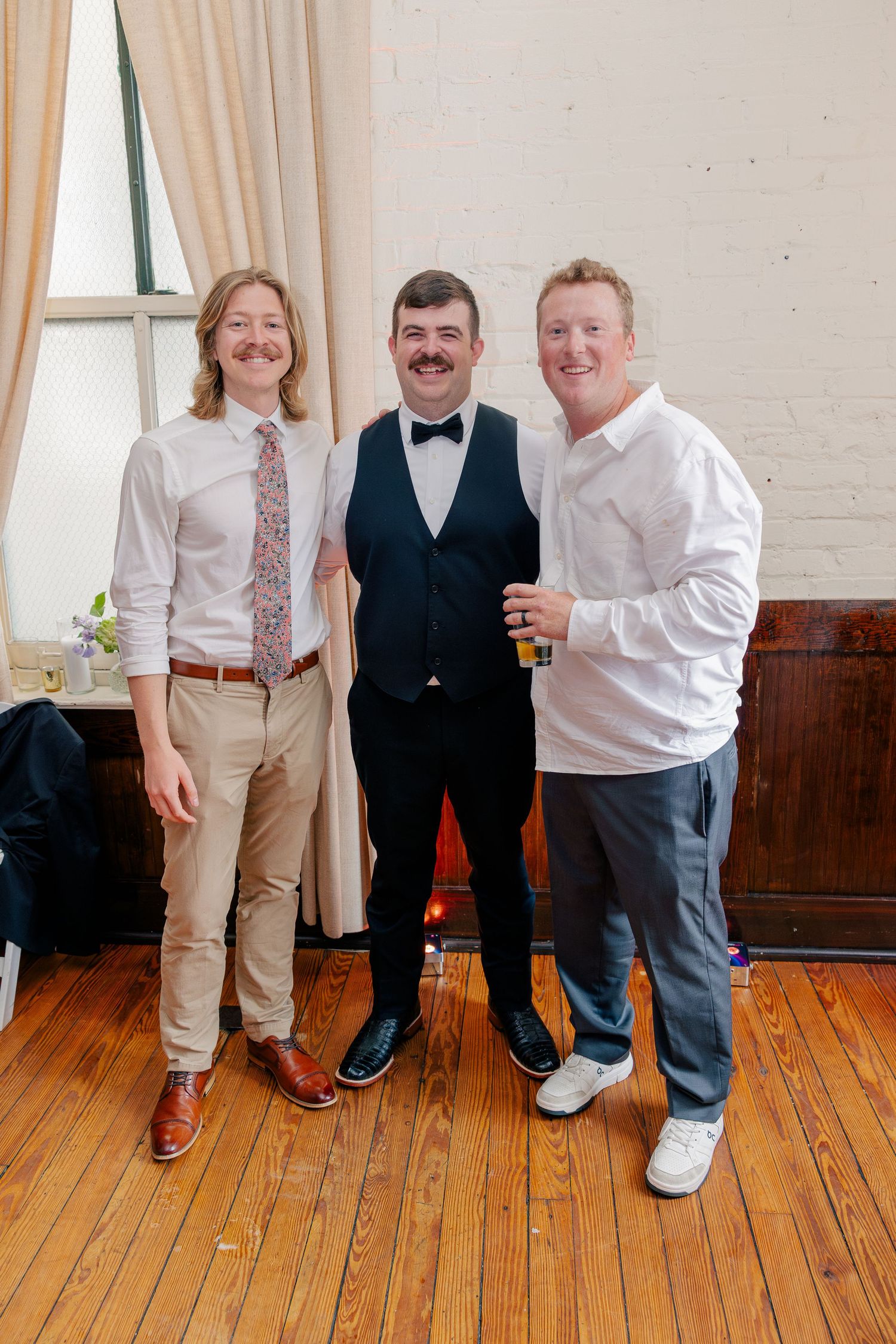 Three men in formal attire smiling together at wedding reception in rustic white brick venue.