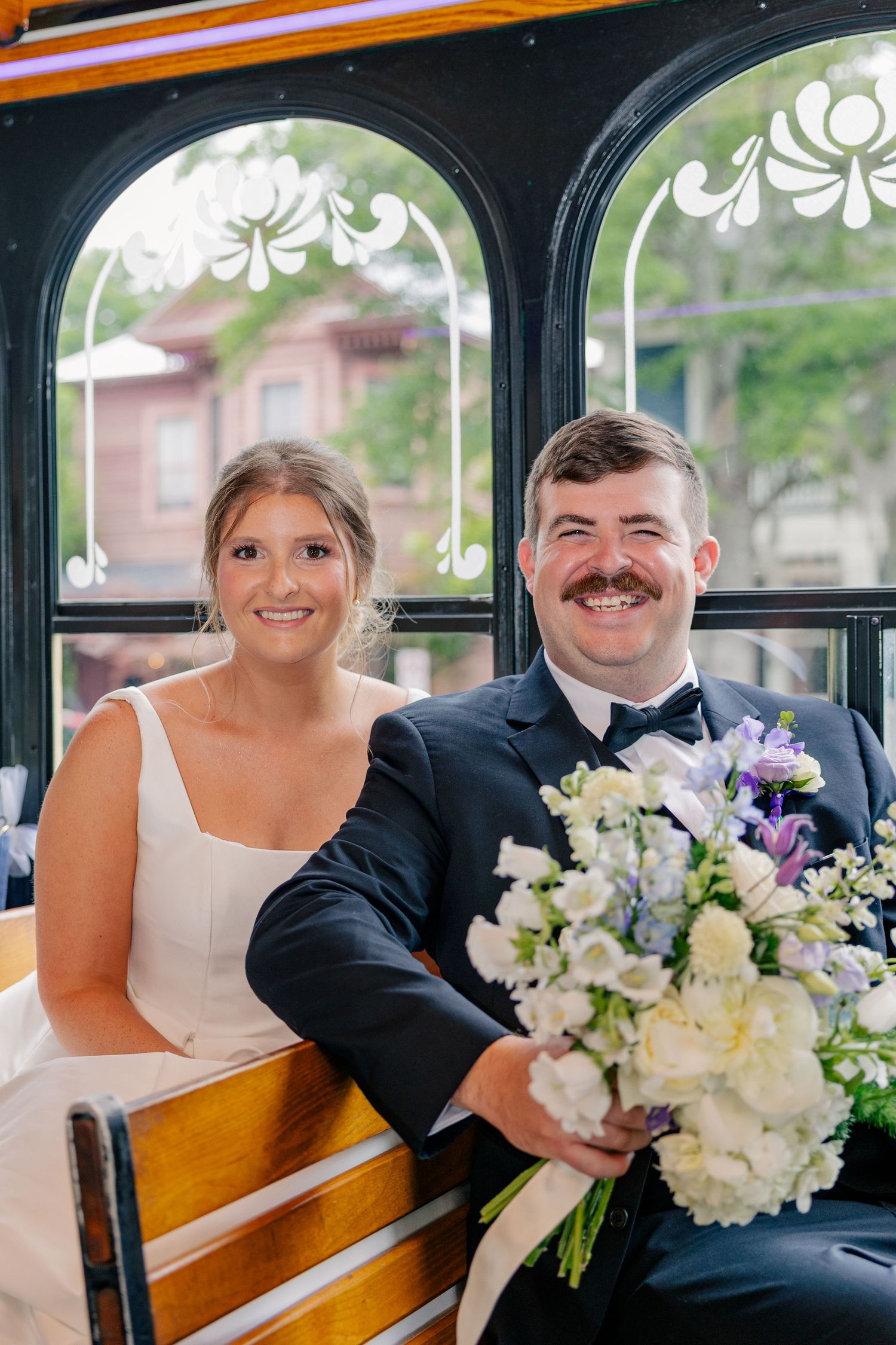 Smiling bride and groom sit together on vintage trolley holding white and purple wedding bouquet.