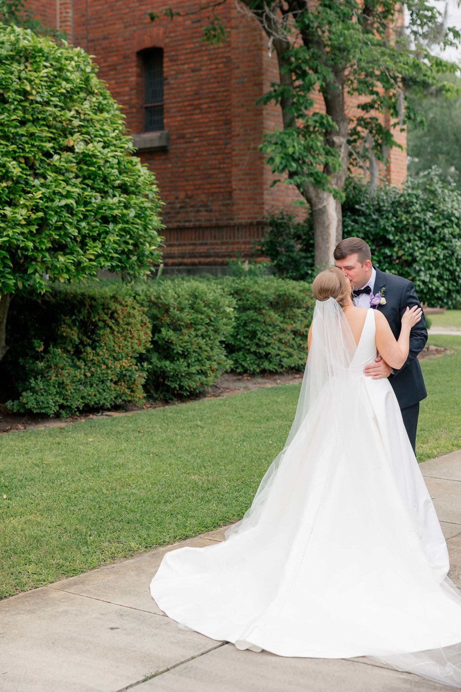Bride and groom share a romantic kiss outdoors near a brick building surrounded by lush green hedges.