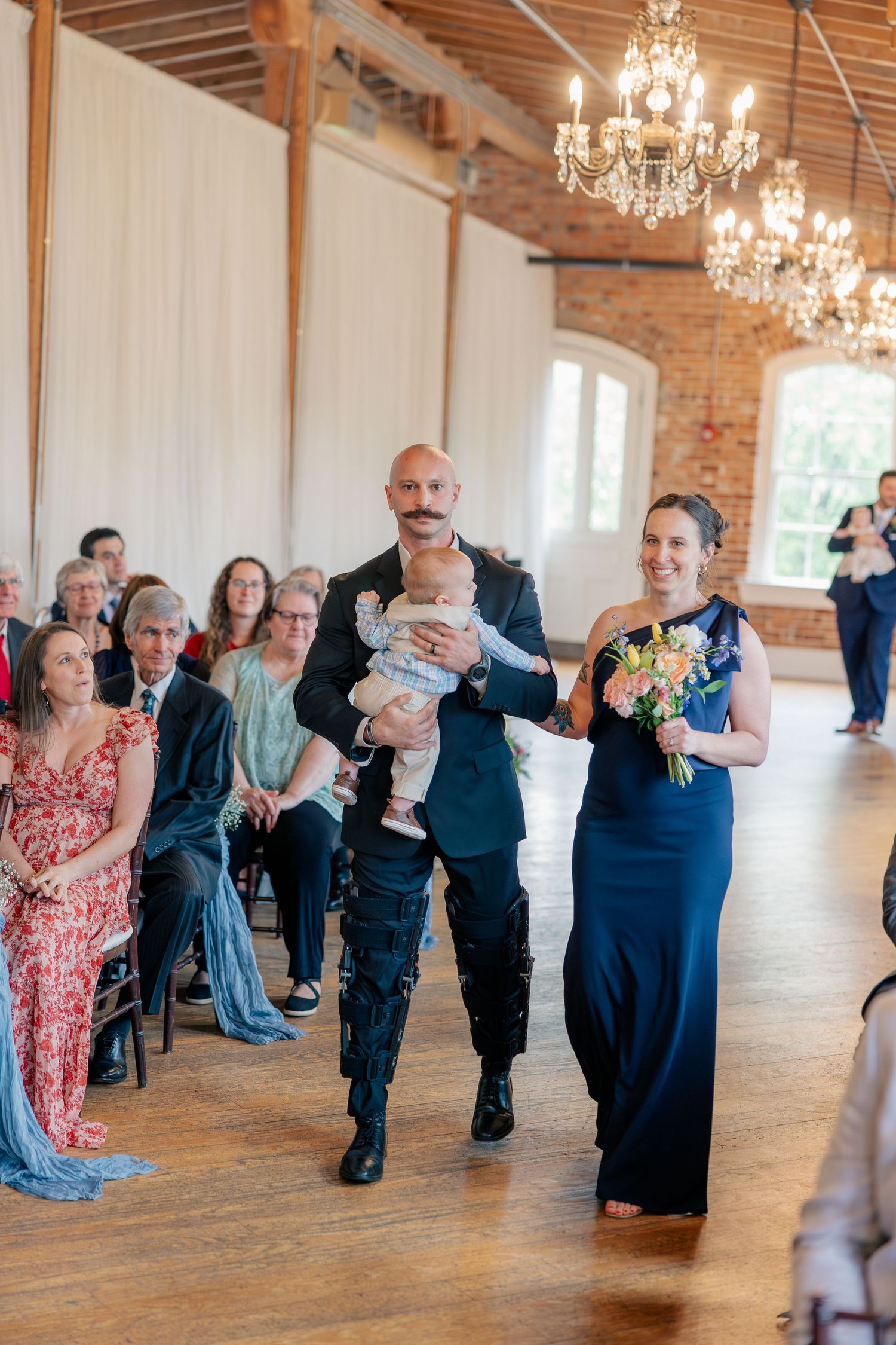 Groomsman carrying a baby walks down the aisle with a bridesmaid in navy dress at an elegant brick venue wedding.