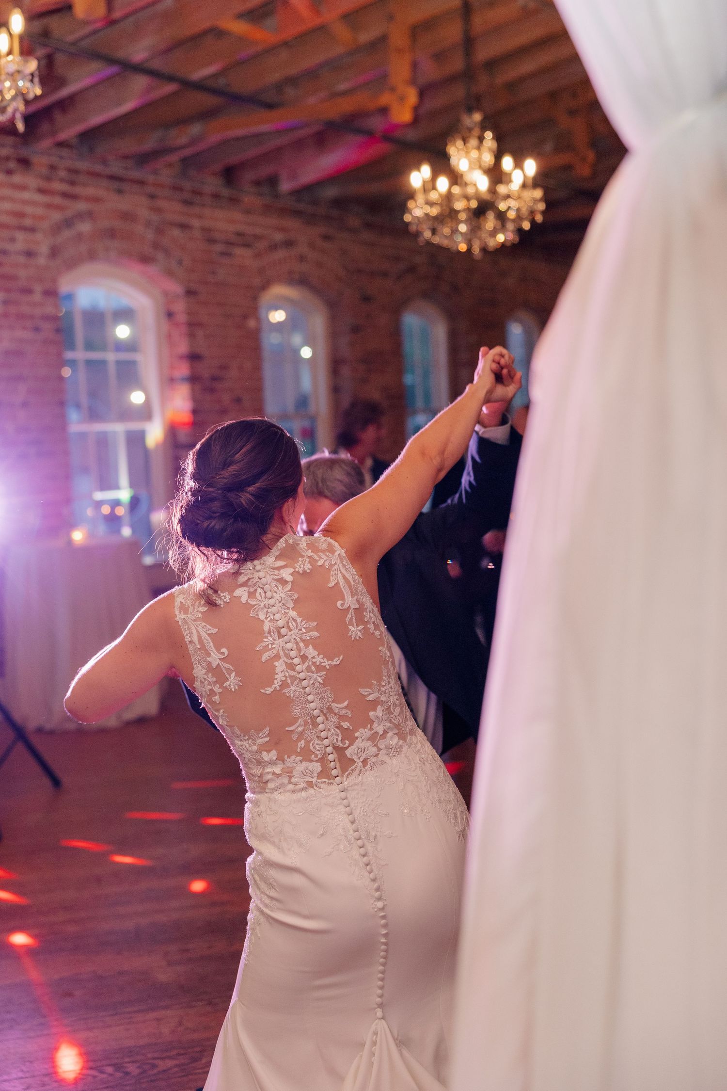 Bride in lace-back wedding gown raises hand while dancing at elegant brick venue reception.