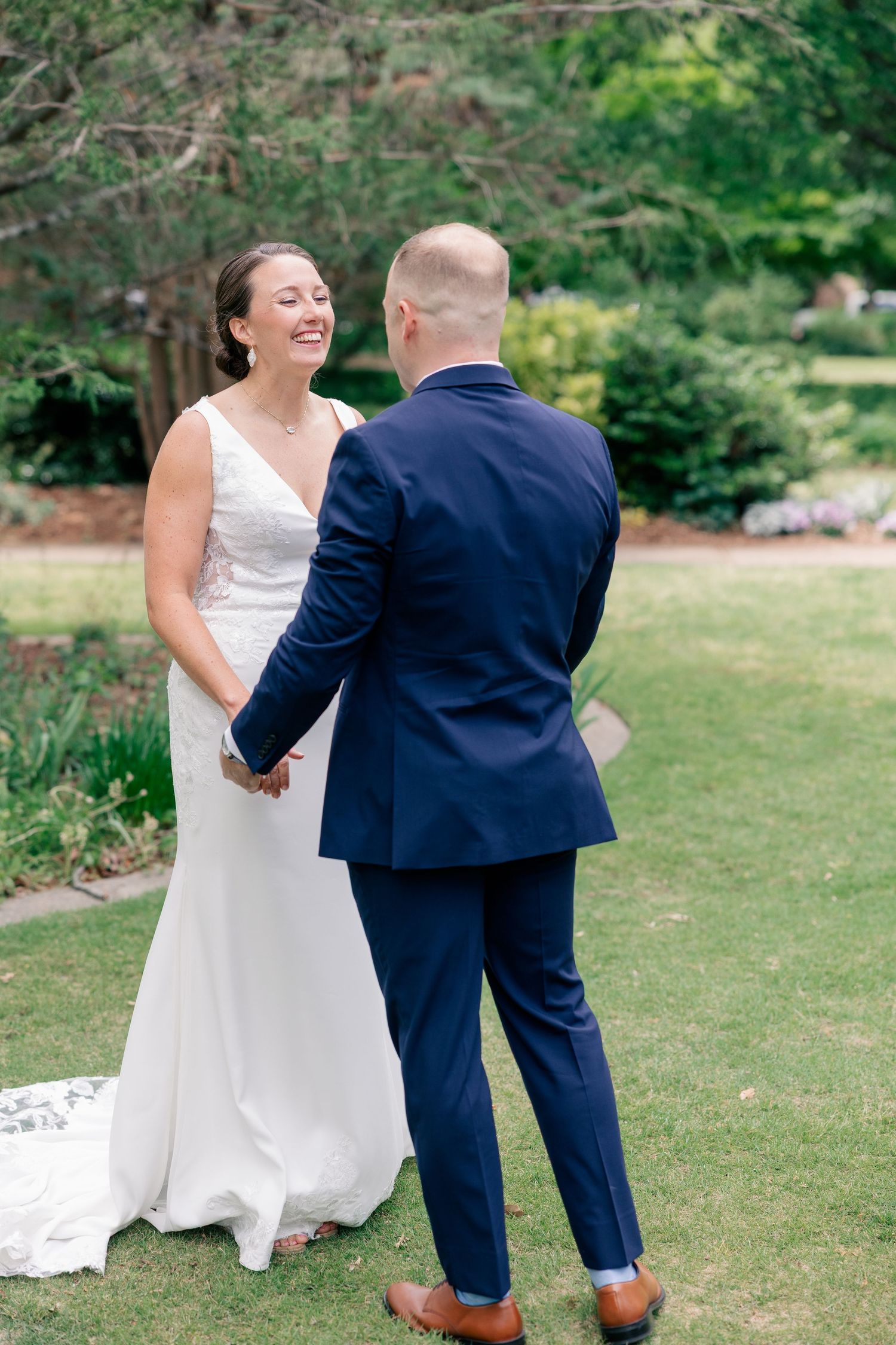 Bride and groom share joyful first look moment, holding hands and laughing in lush green garden setting.