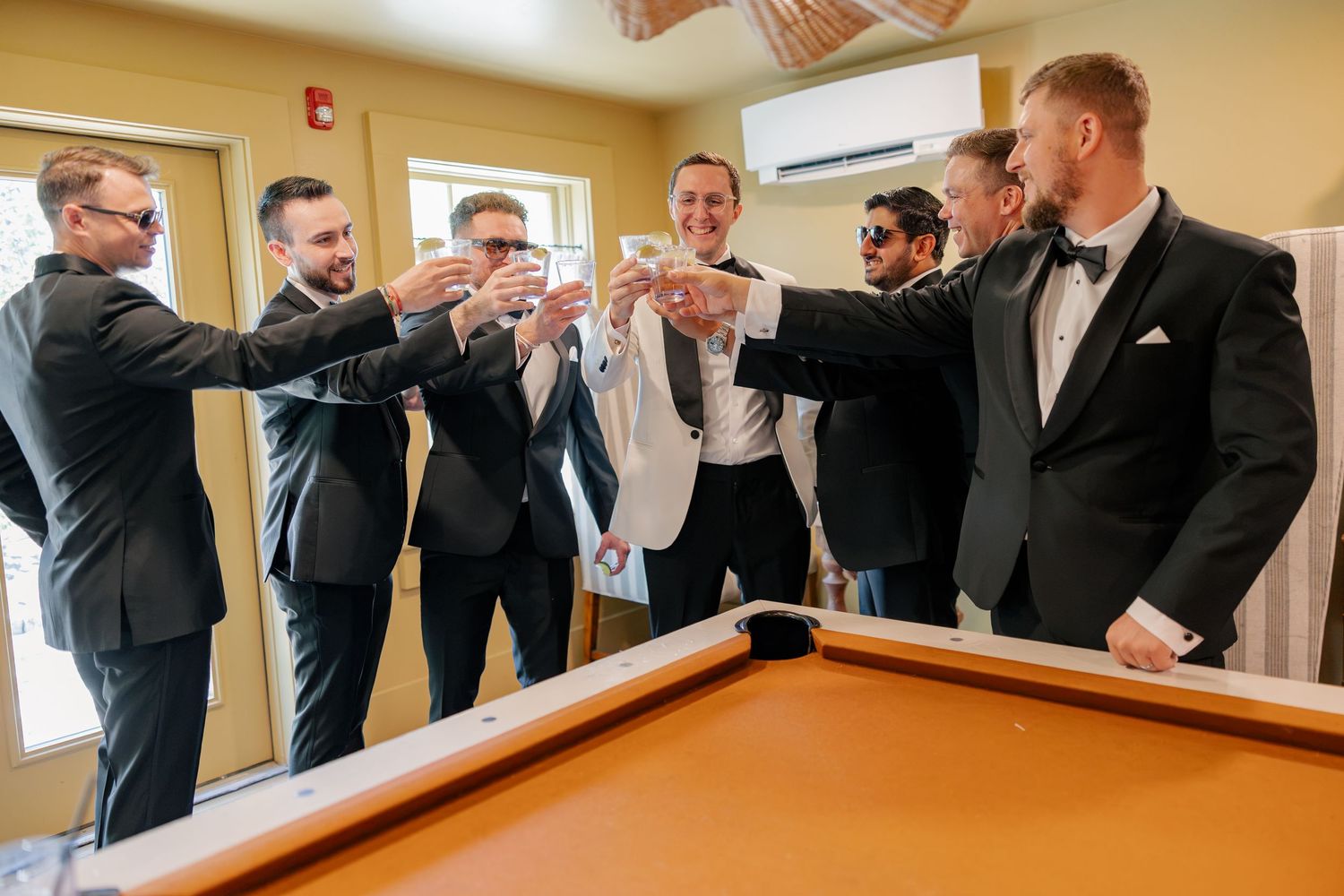 Groom and groomsmen toast together in tuxedos beside a pool table before wedding ceremony.