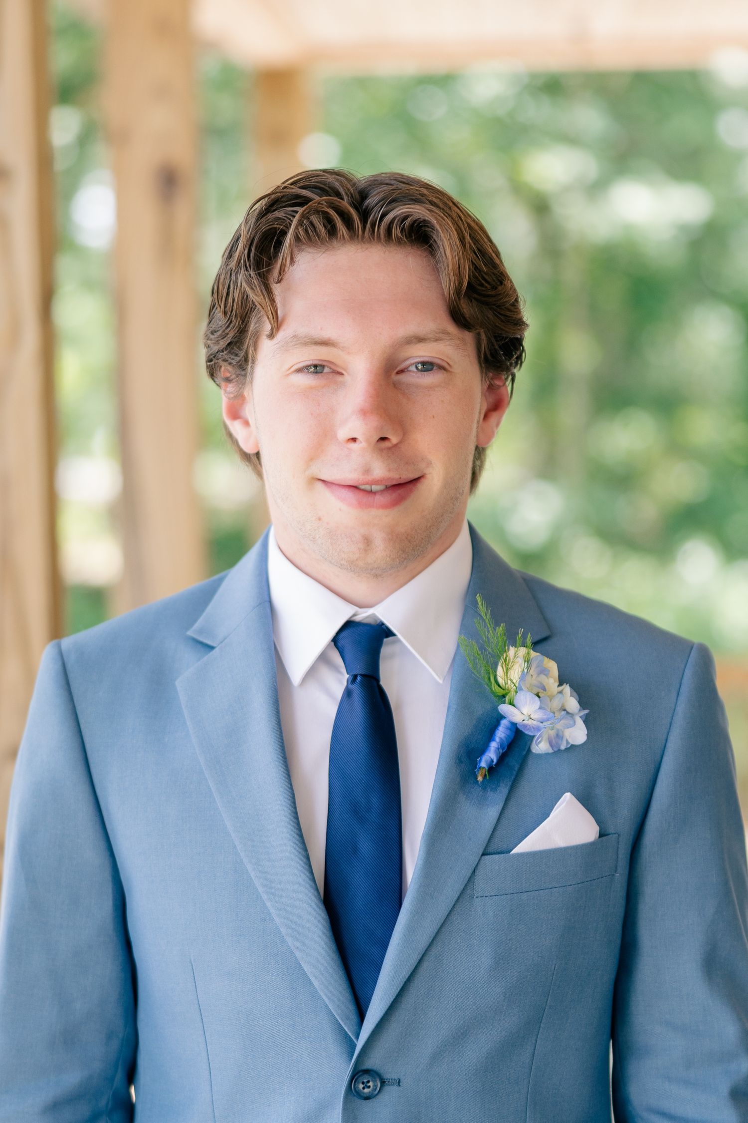 Handsome groom in light blue suit and navy tie with boutonniere smiles for a portrait at his wedding.