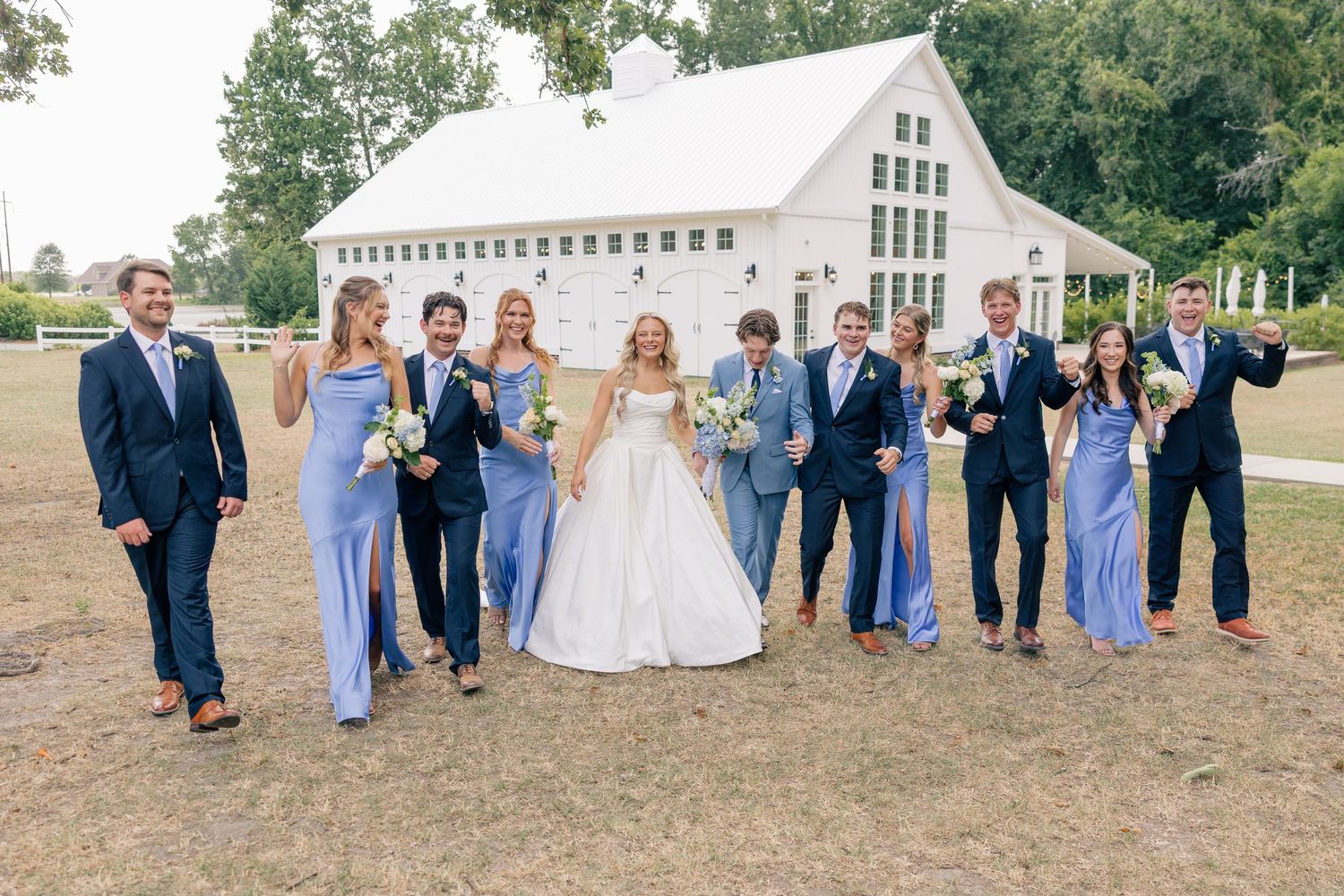 Wedding party in blue attire posing in front of a white barn venue on a sunny day.