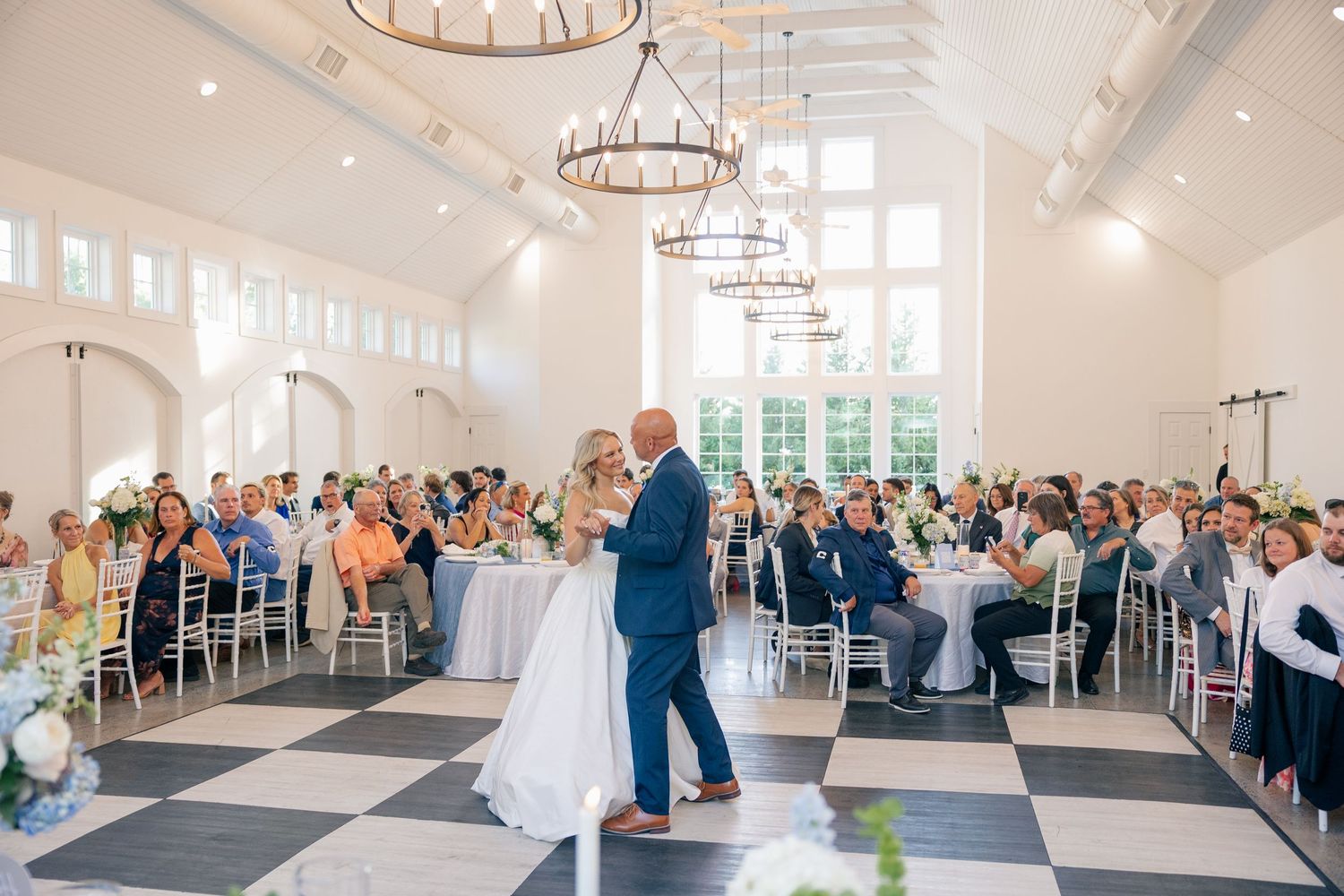 Bride and groom share first dance in elegant white ballroom with checkered floors and chandelier lighting.